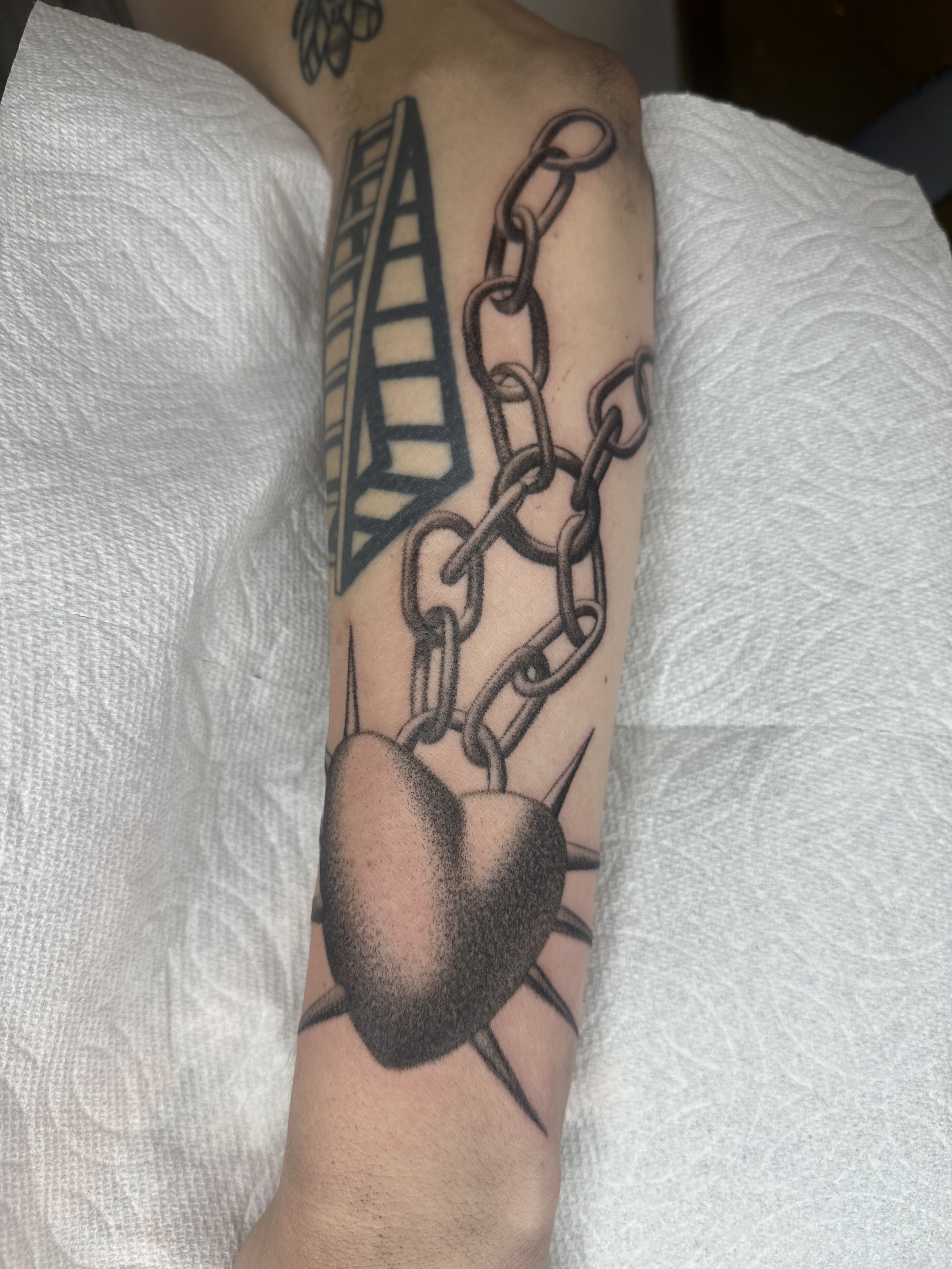 Tattoo of a chain with a heart-shaped lock on a person's arm, with a geometric pyramid design above.