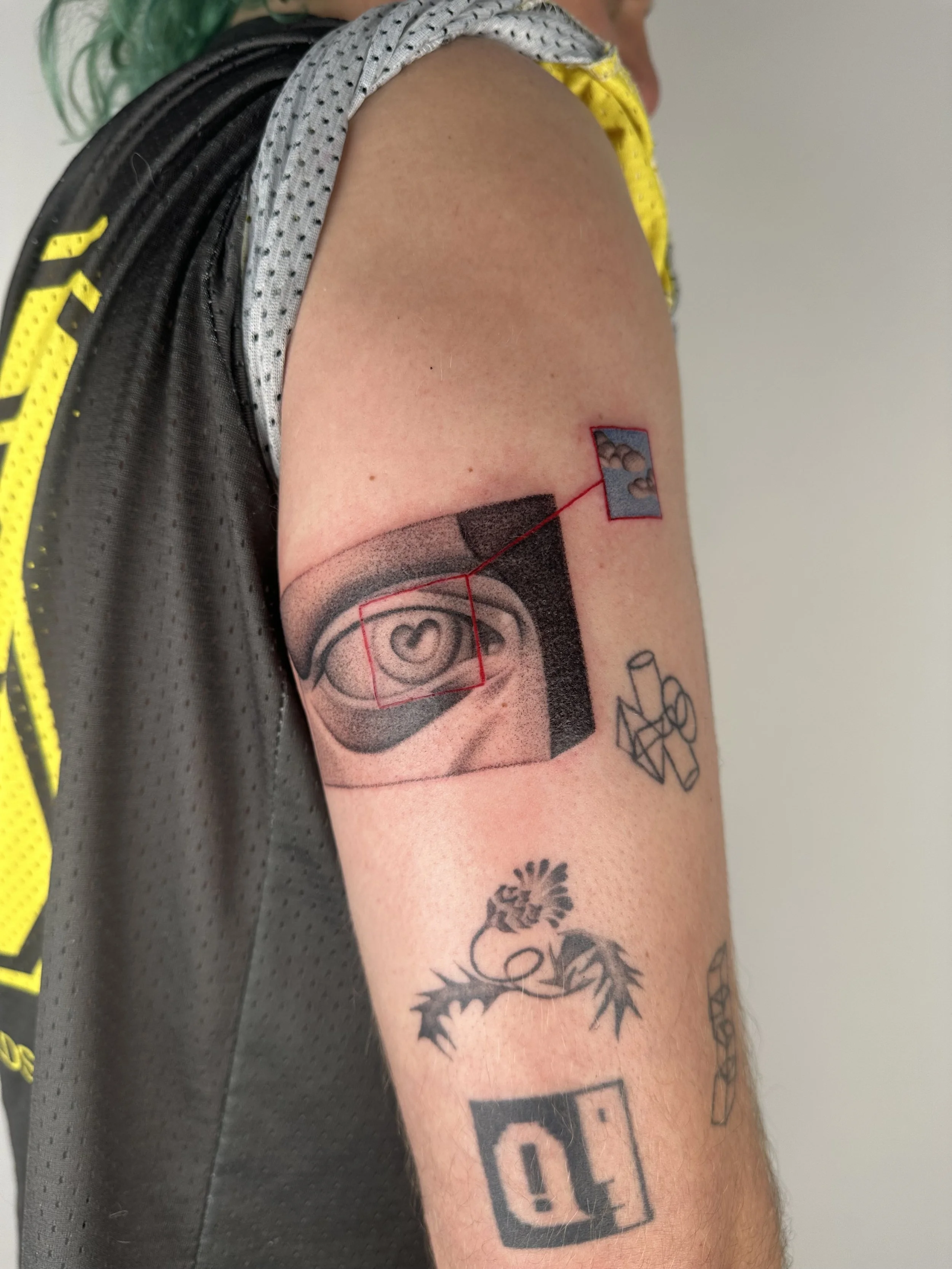 Tattoo of an eye with a heart in the center on a person's upper arm, with geometric design and small reference image connected by red lines, and two additional tattoos of a dragon and an abstract symbol below.