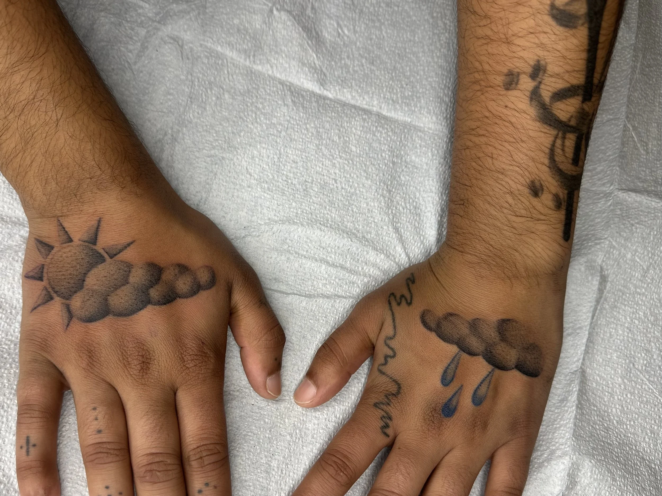 Two hands with tattoo illustrations, one with a 3D rendered sun and cloud tattoo, the other with a cloud and rain tattoo, resting on a white surface.
