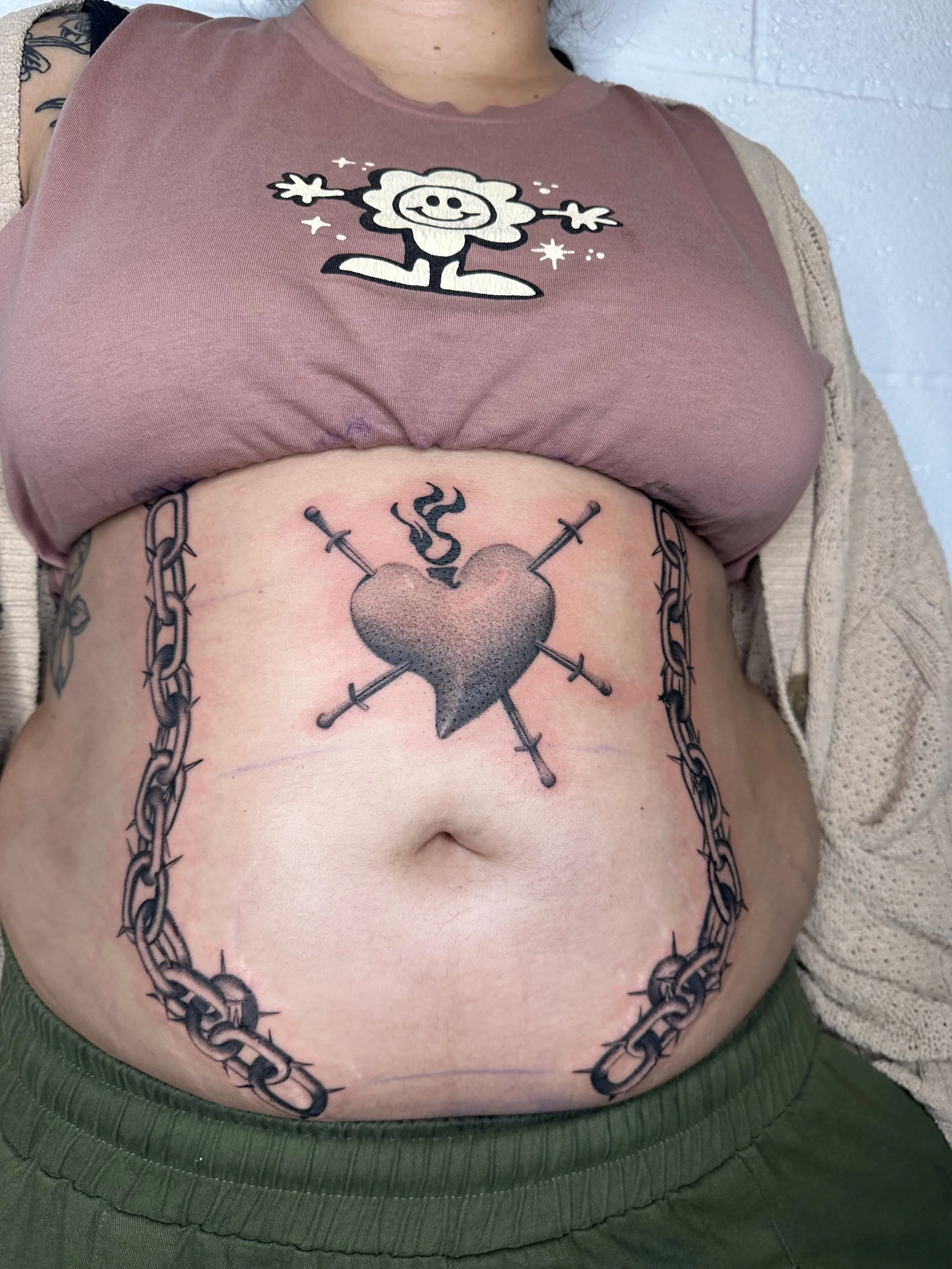 Tattoo of a heart with three swords piercing it and a small flame or smoke above it, surrounded by a chain border on the stomach.