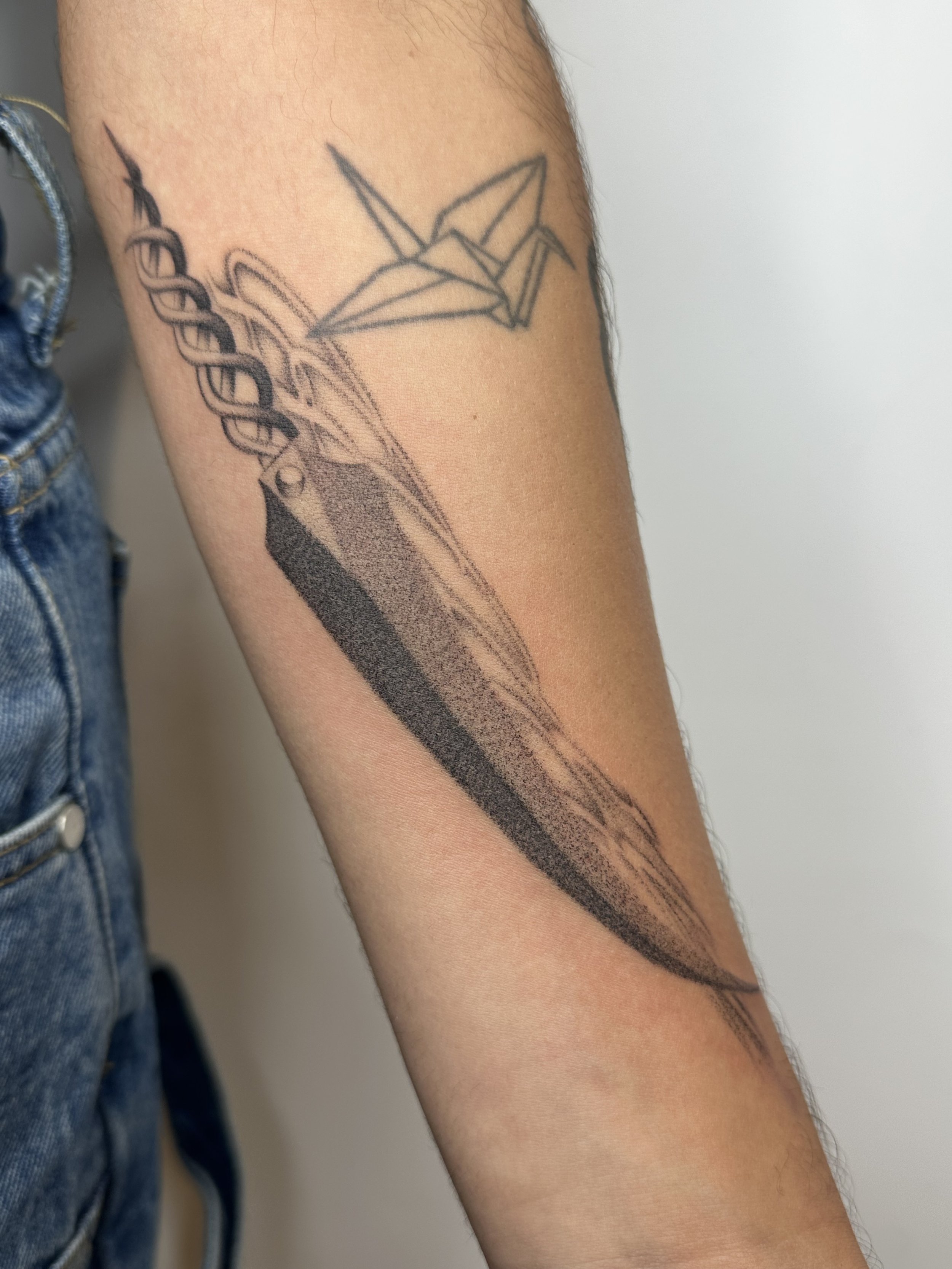 Tattoo of a paper boat origami and a stylized sword with a chain design on a person's forearm.