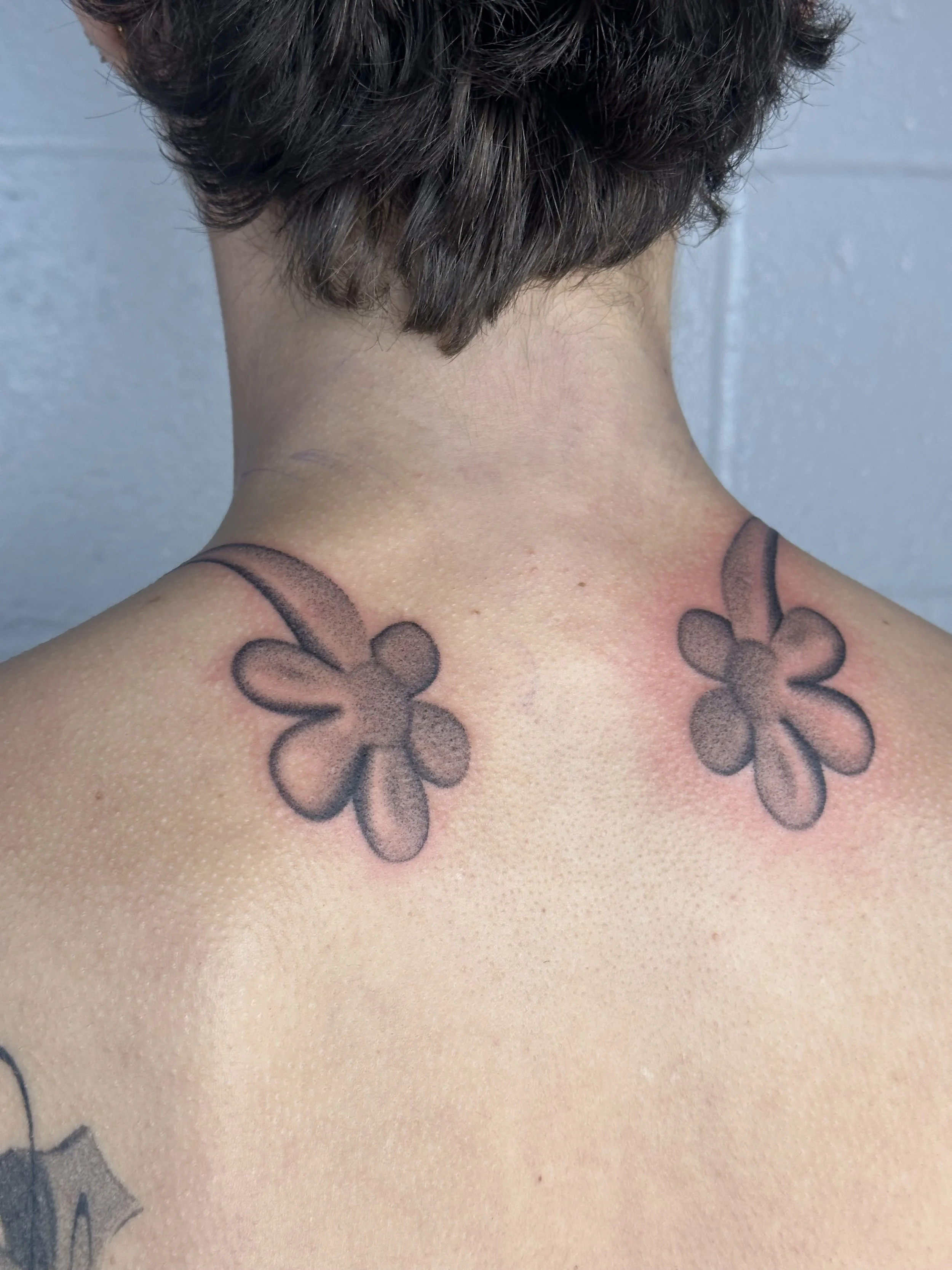 Close-up view of a person's upper back and neck showing two tattoos of flowers with black outlines and shading, placed on each side of the neck.