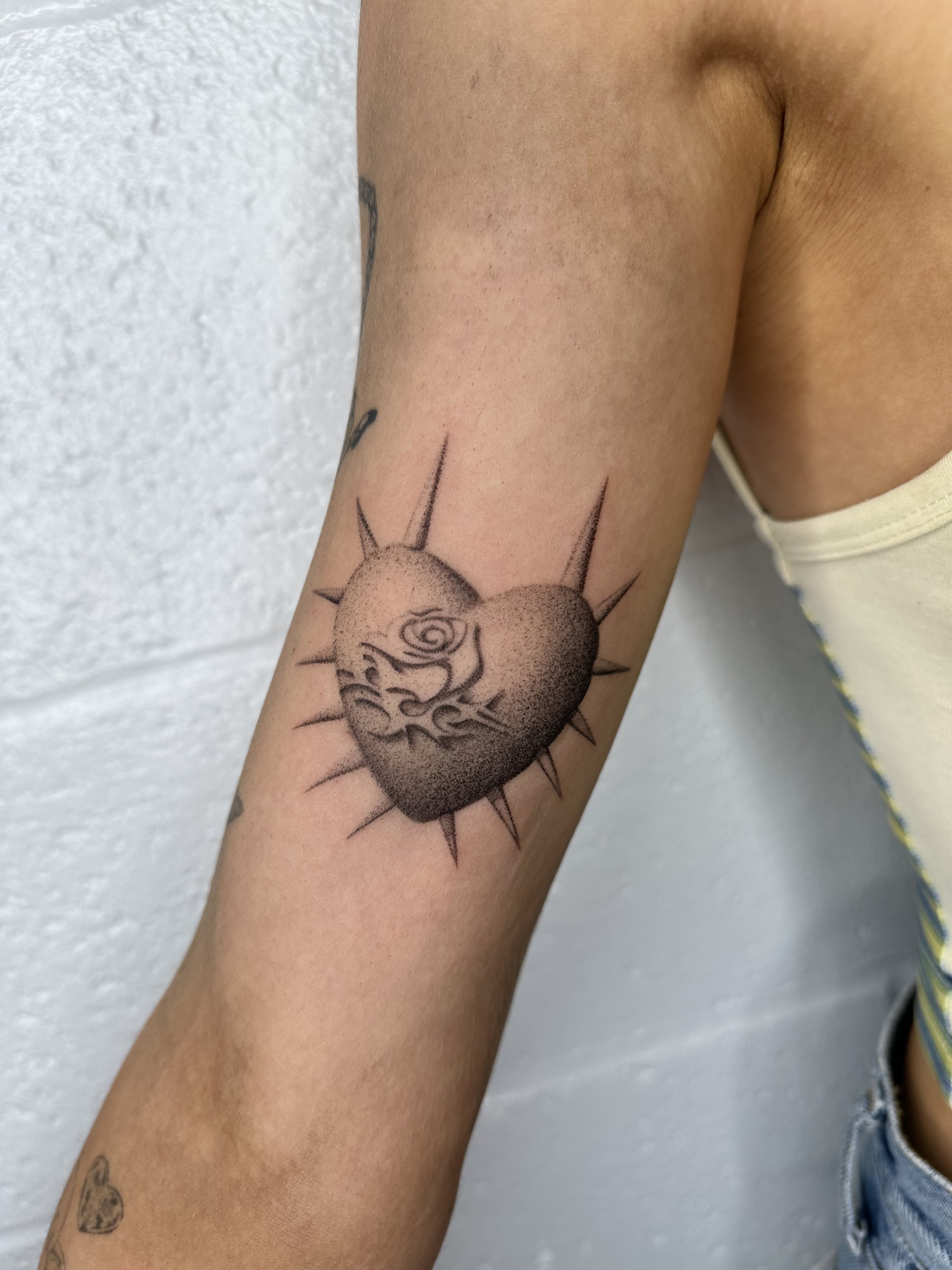 Tattoo of a heart with a face and flames, surrounded by rays, on a person's arm.