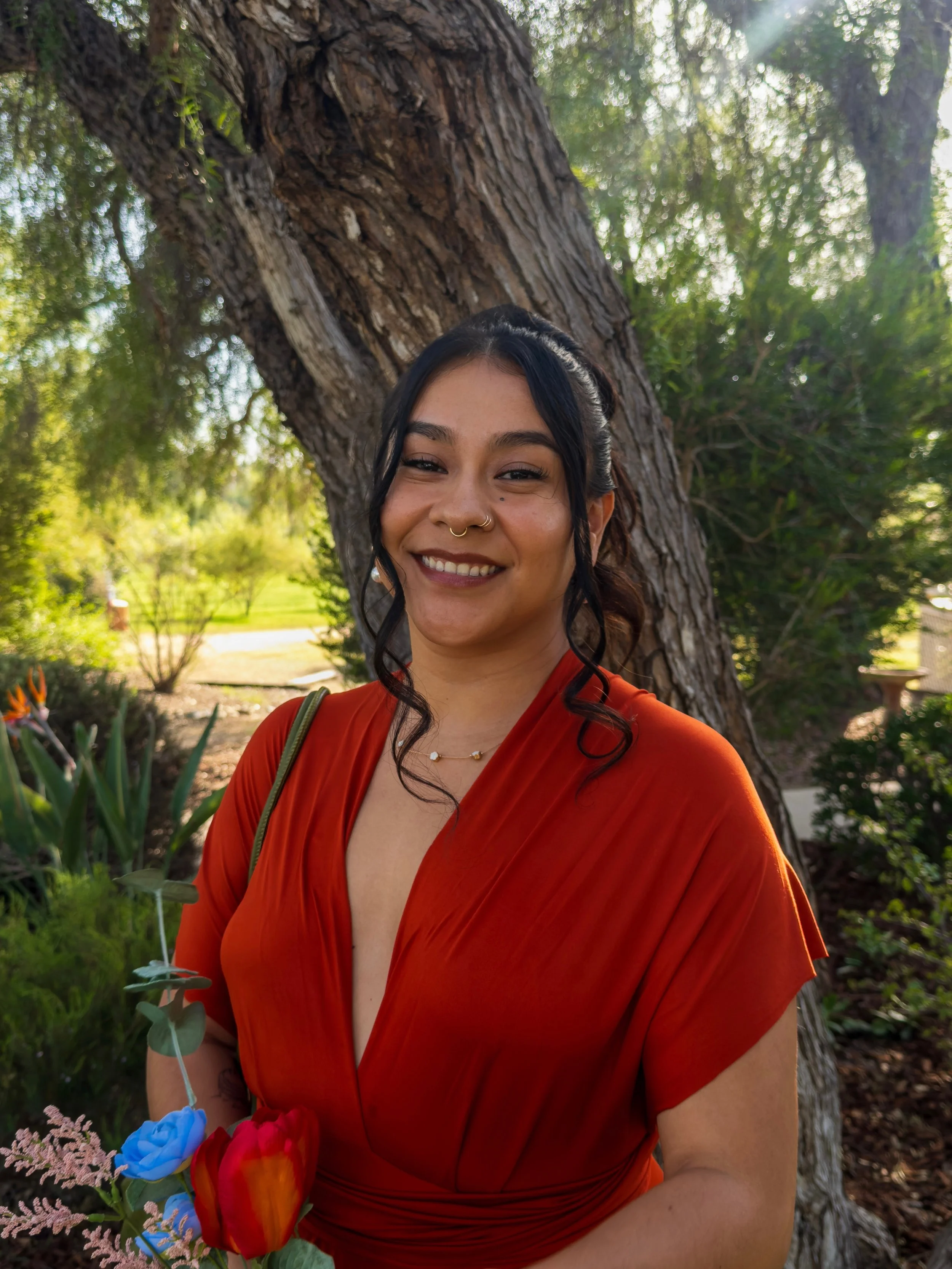 A smiling woman with dark hair, wearing a red dress, stands outdoors near a large tree with sunlight filtering through the leaves.