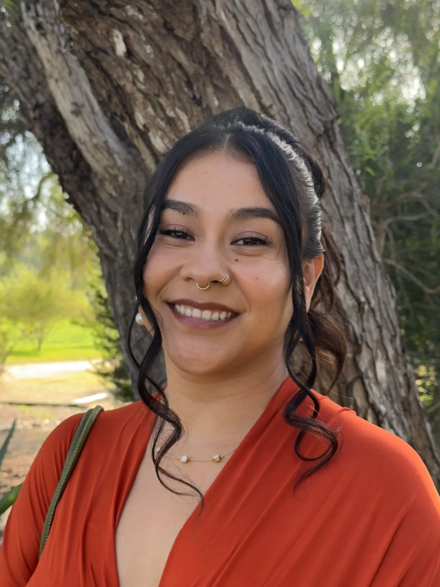 A young woman with dark hair and tattoos, smiling outdoors in front of a large tree, wearing an orange top and a delicate necklace.