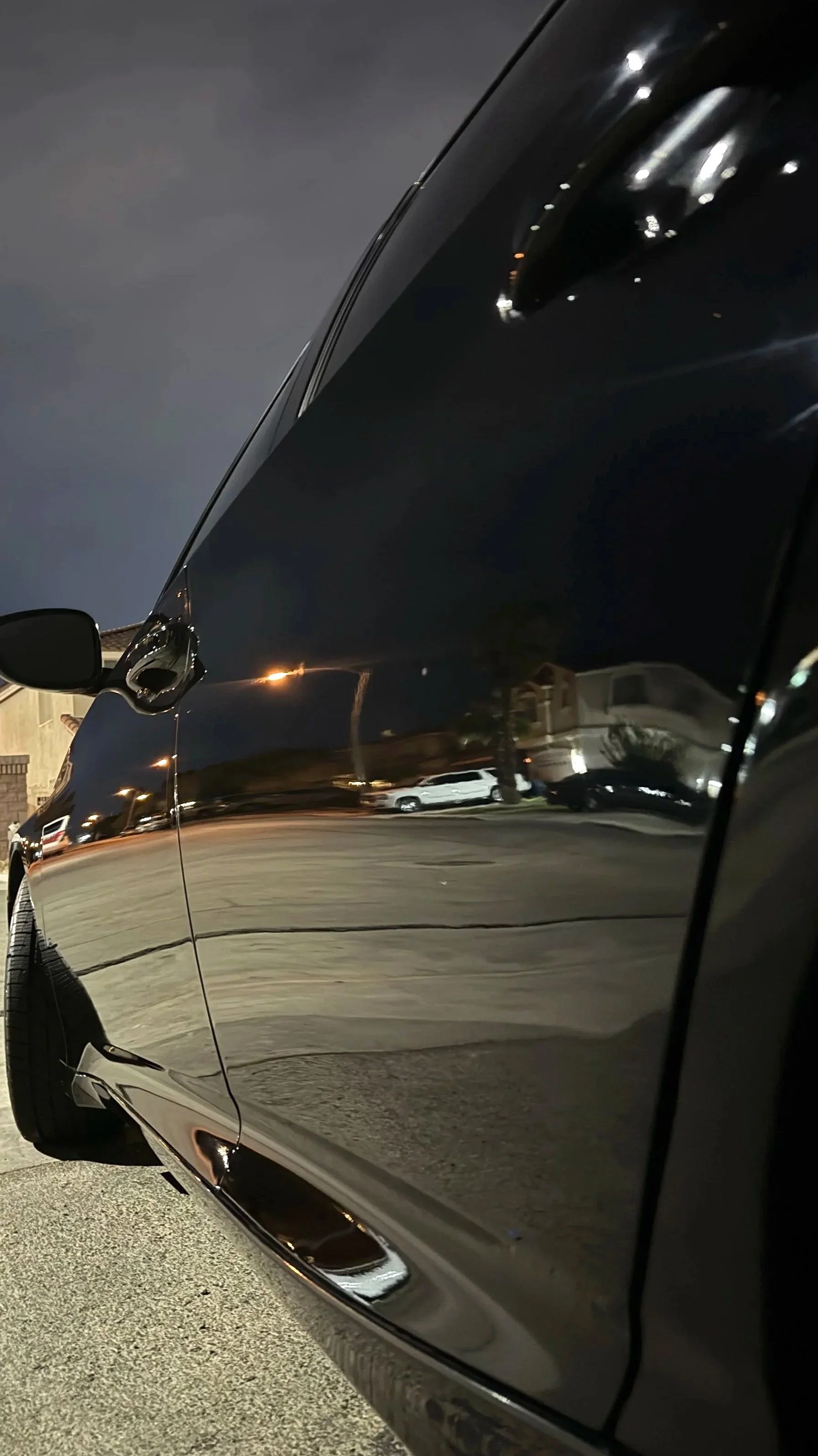 Close-up of a shiny black car at night, reflecting the streets and houses around it.
