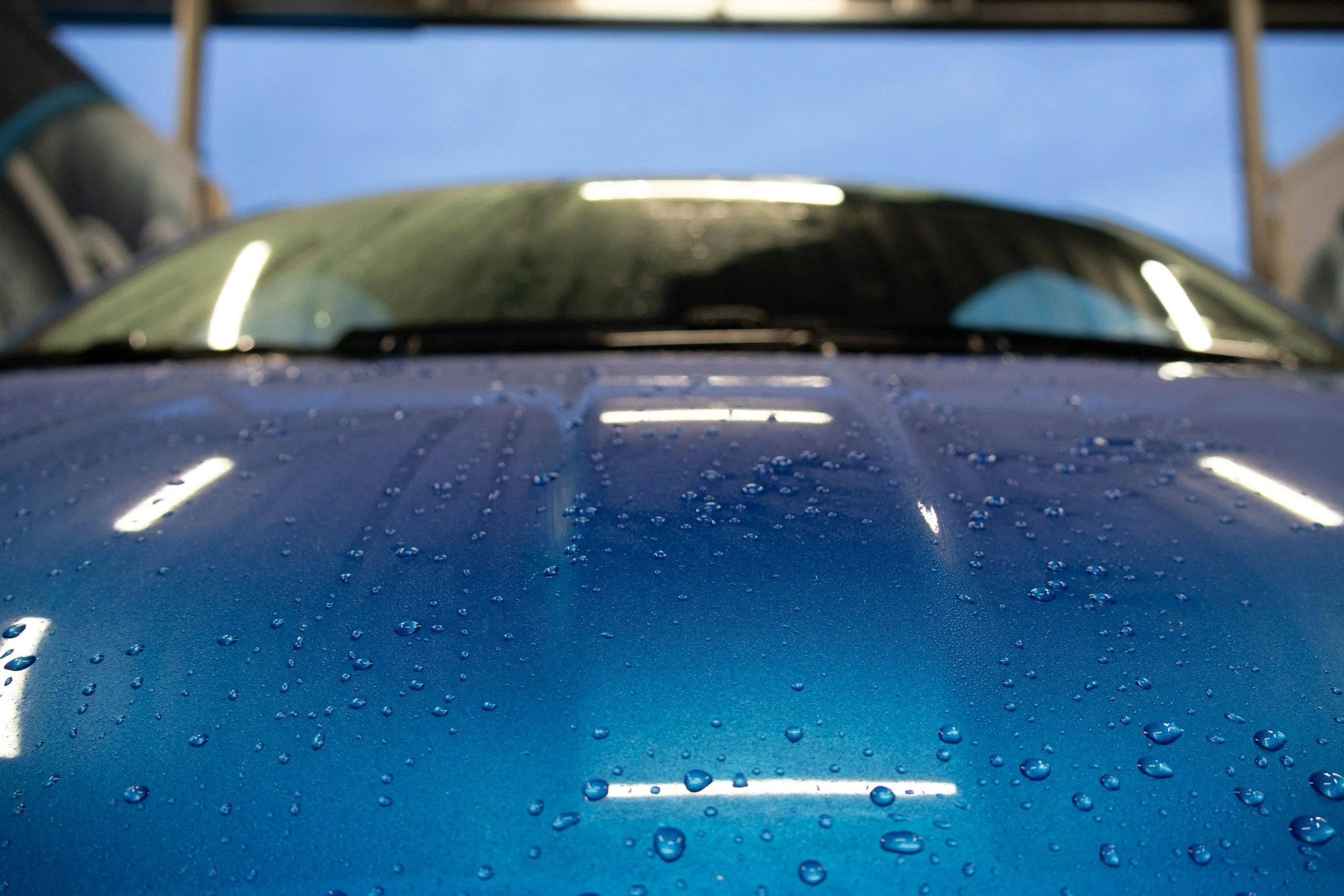Close-up of a blue car's hood with water droplets and the windshield in the background.
