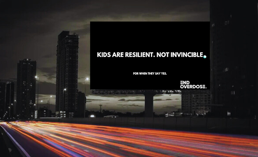 A billboard against a dark cityscape at night displaying the message 'Kids are resilient. Not invincible. For when they say yes. End overdose.'