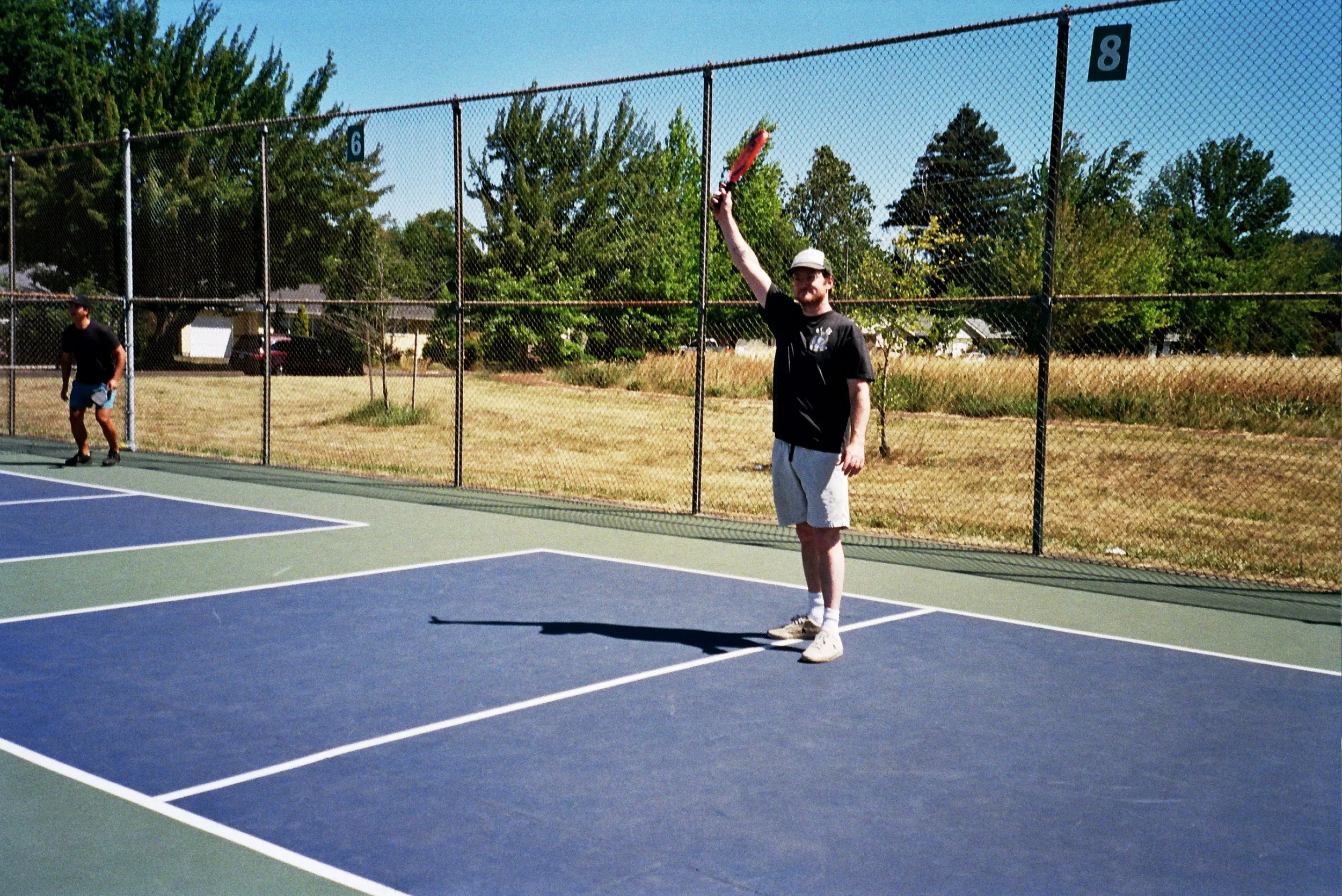A man in a black T-shirt, shorts, and a hat holds a red pickleball paddle on a tennis court, with another person in the background near the fence, under a clear blue sky and surrounded by trees.