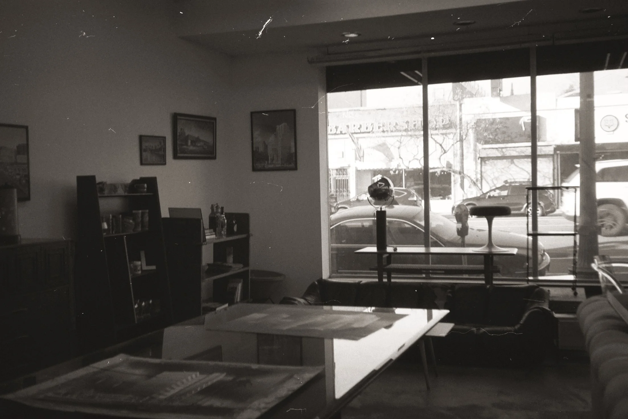 A black and white photo of the interior of a cafe or bakery, with a large window showing parked cars and buildings outside. Inside, there are framed pictures on the wall, tables, chairs, a bookshelf, and a globe on a stand near the window.