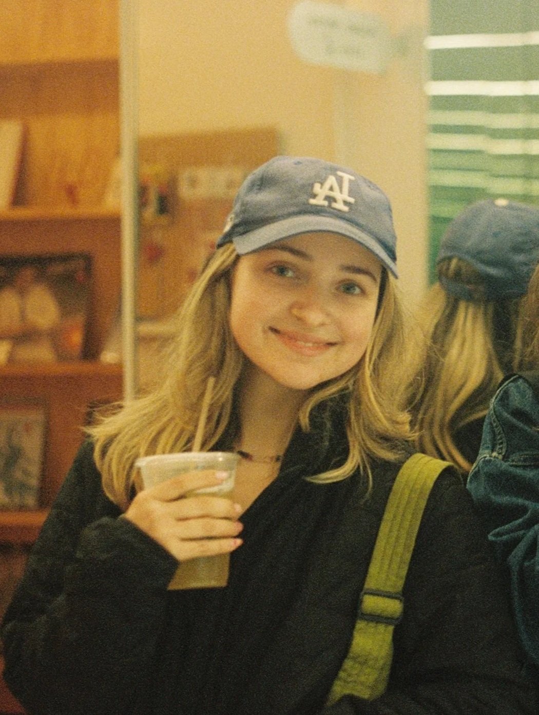 A young woman with blonde hair wearing a blue Los Angeles Dodgers baseball cap, smiling at the camera, holding a drink in a plastic cup, and carrying a green backpack, in a cozy indoor setting.