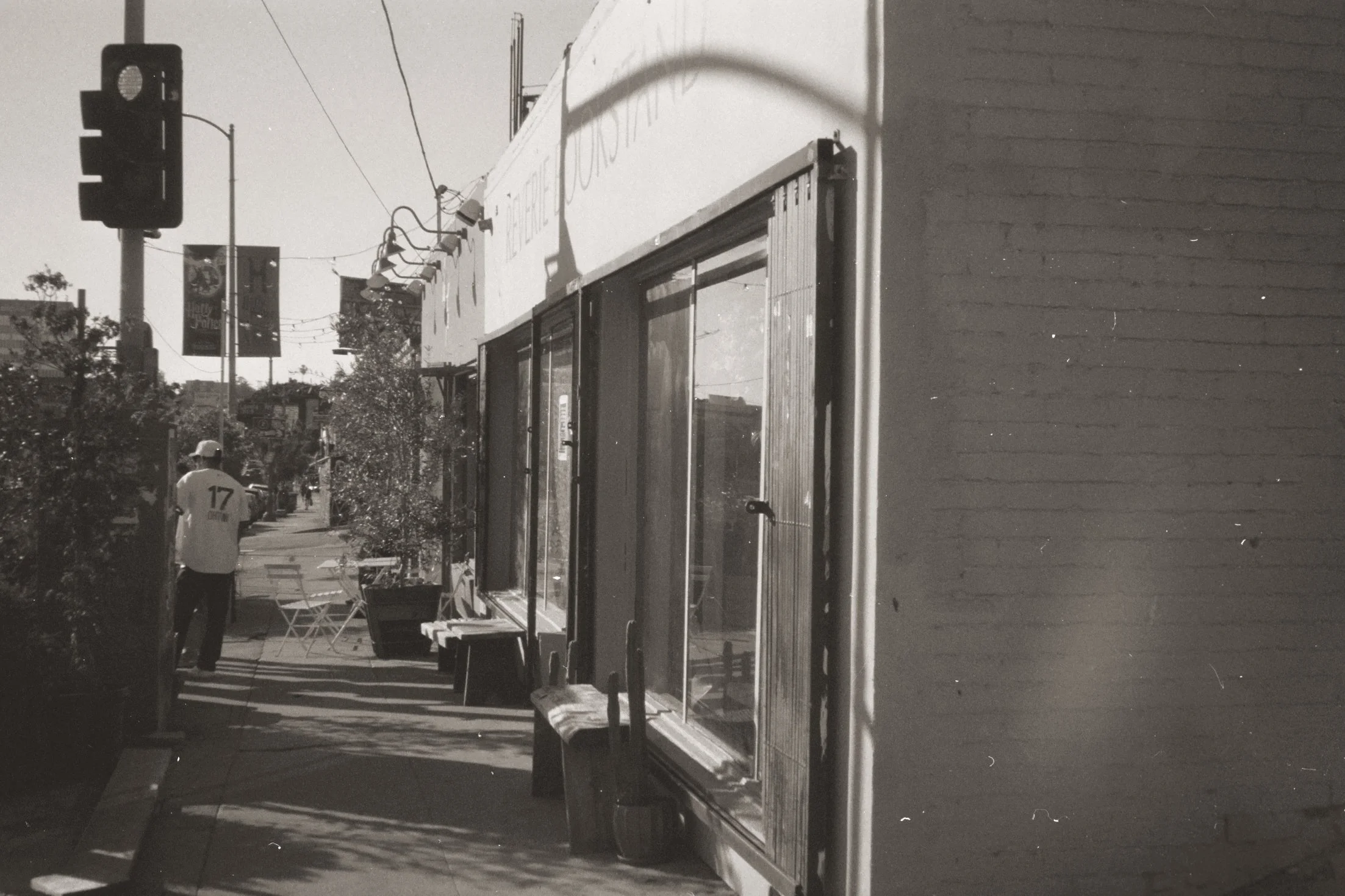 Black and white photo of a city sidewalk scene, featuring a building with large windows, a lamppost, and a traffic light. A person wearing a baseball cap and a jersey with the number 17 on the back walks along the sidewalk.