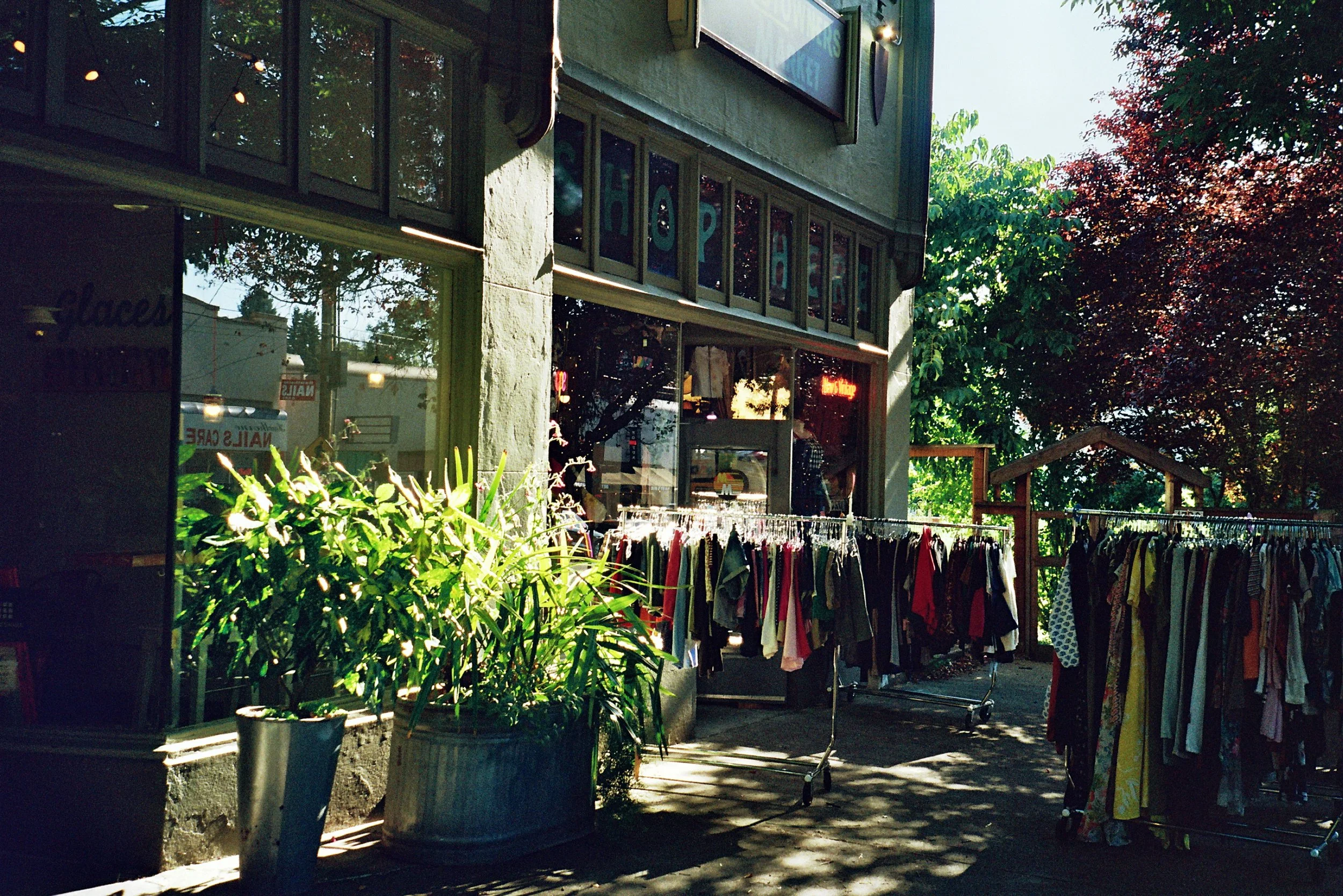 An outdoor clothing store with racks of clothes, plants, and a building with large windows and colorful signs in the windows, surrounded by trees and sunlight.
