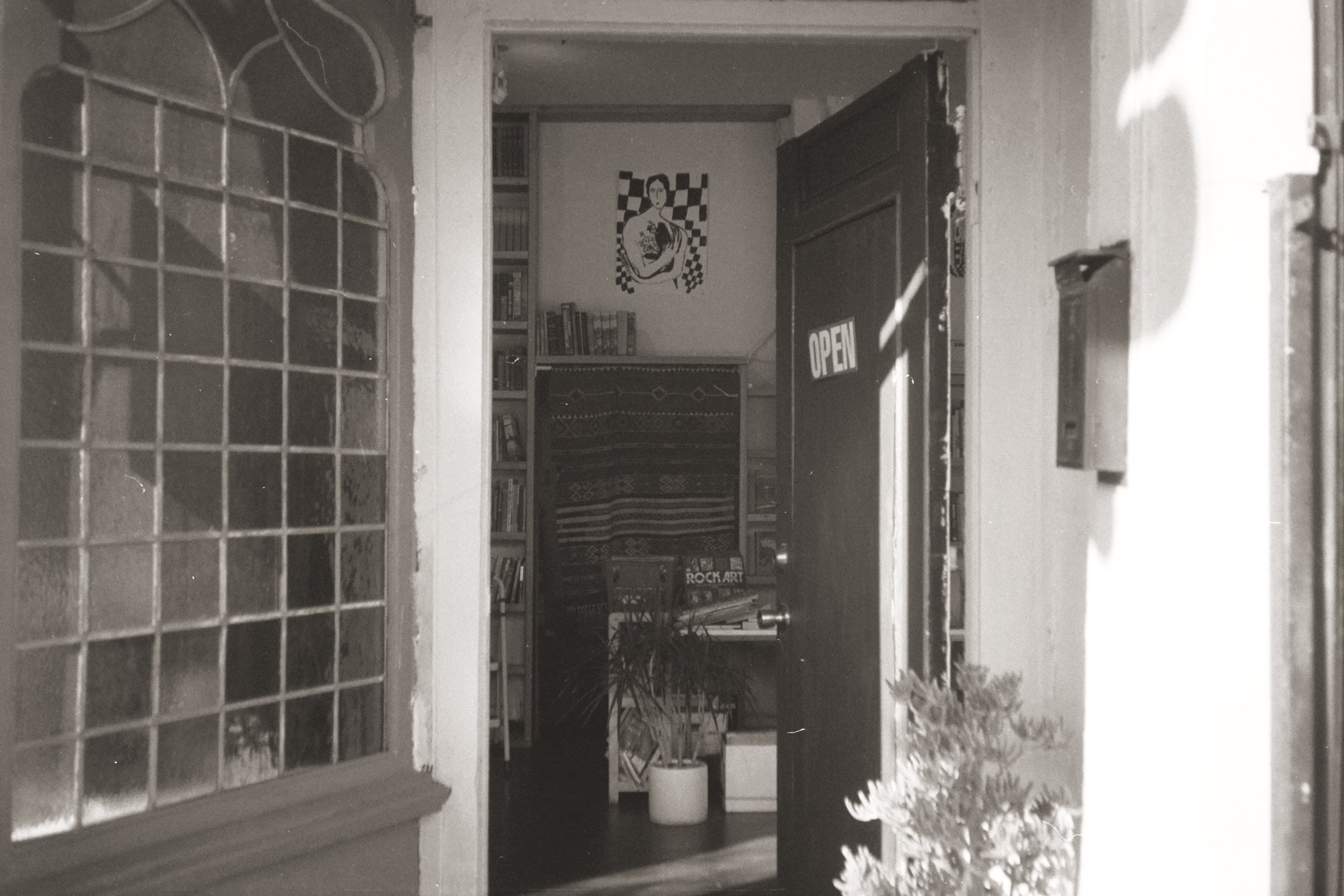 Black and white photo of the entrance to a house with an open door. The door has a sign saying 'OPEN' and the house interior is visible with bookshelves, a poster on the wall, and various items, including a potted plant and a patterned cloth.