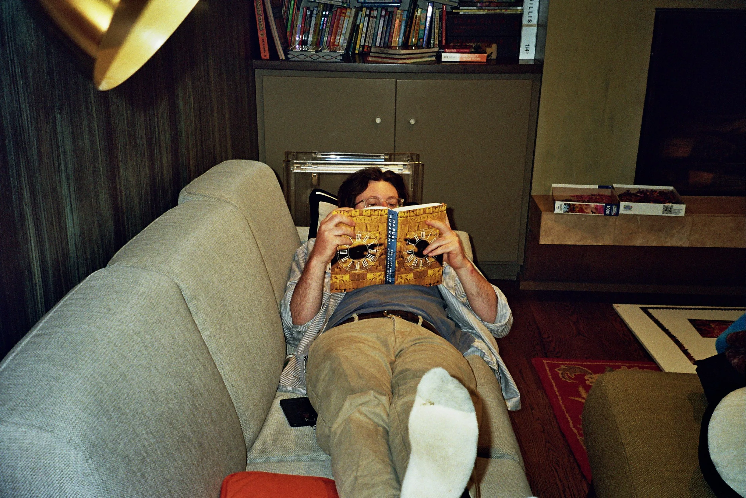 A man with brown hair and glasses lying on a beige couch, reading a book with a yellow brick pattern and clock numbers on the cover. He is wearing a striped shirt and khaki shorts, with white socks and white shoes. The room has a dark wood floor, a r