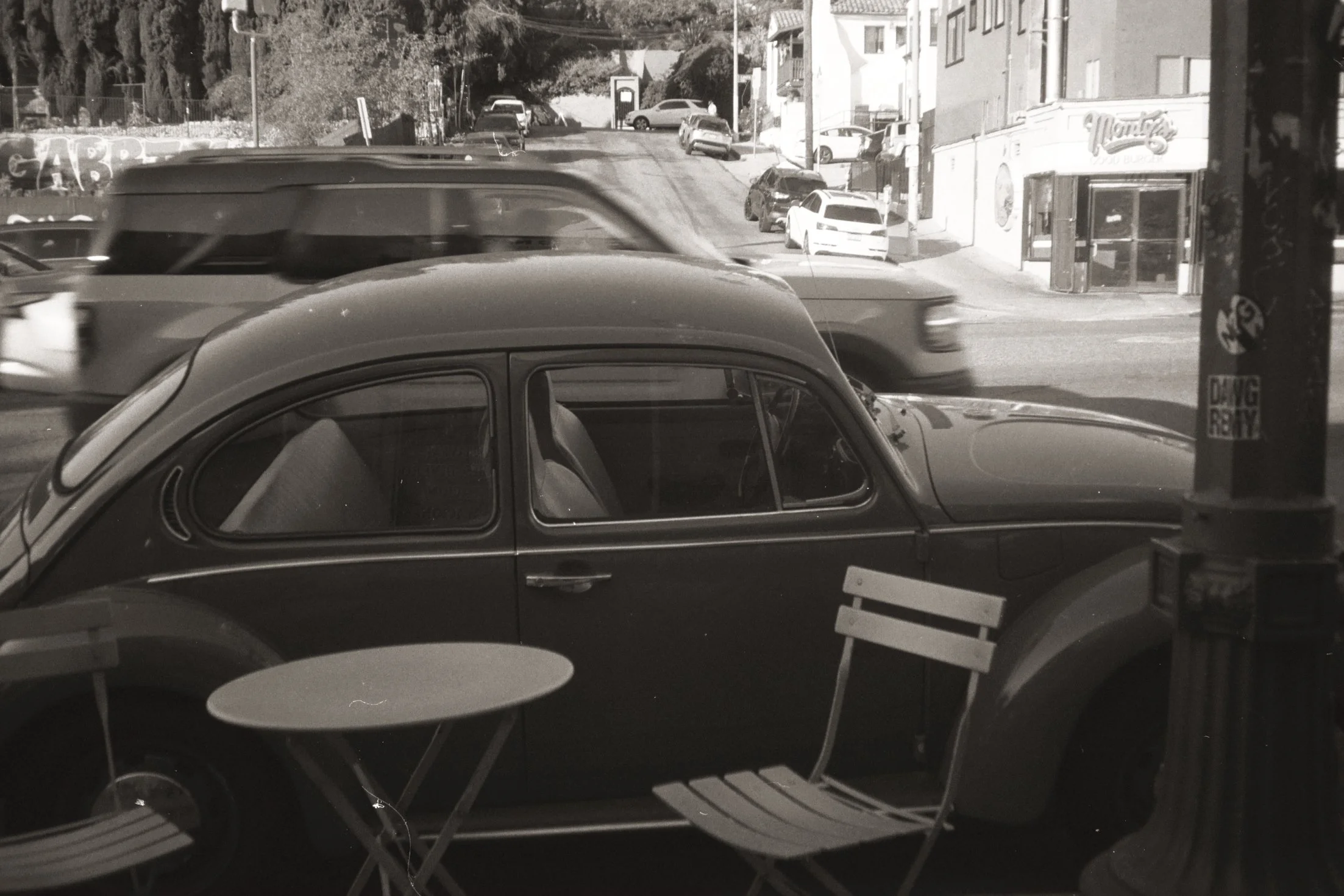 Black vintage car parked outside a cafe with two round tables and chairs, a street scene with multiple cars, a business with a sign and high-rise buildings in the background.