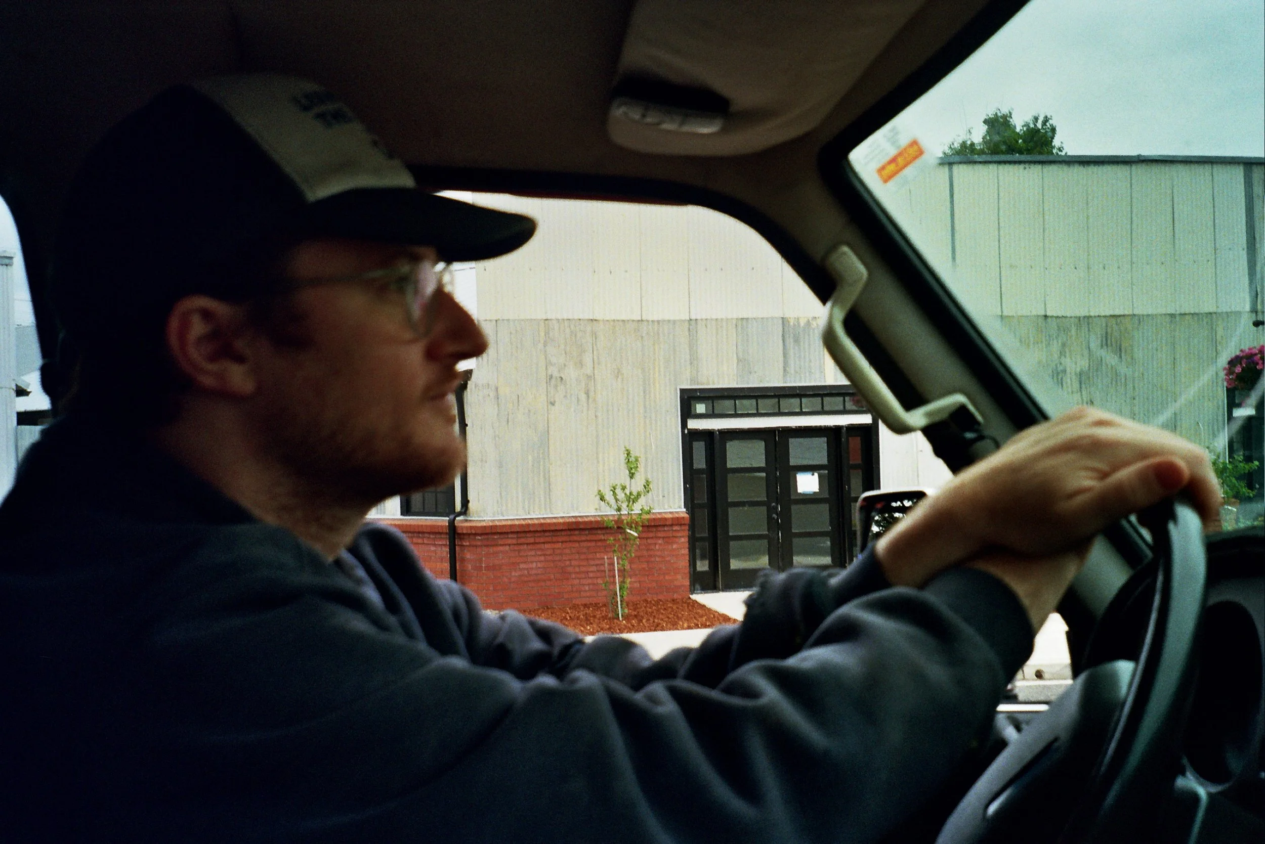 A man with glasses and a cap driving a vehicle, with a building and some greenery visible through the window.