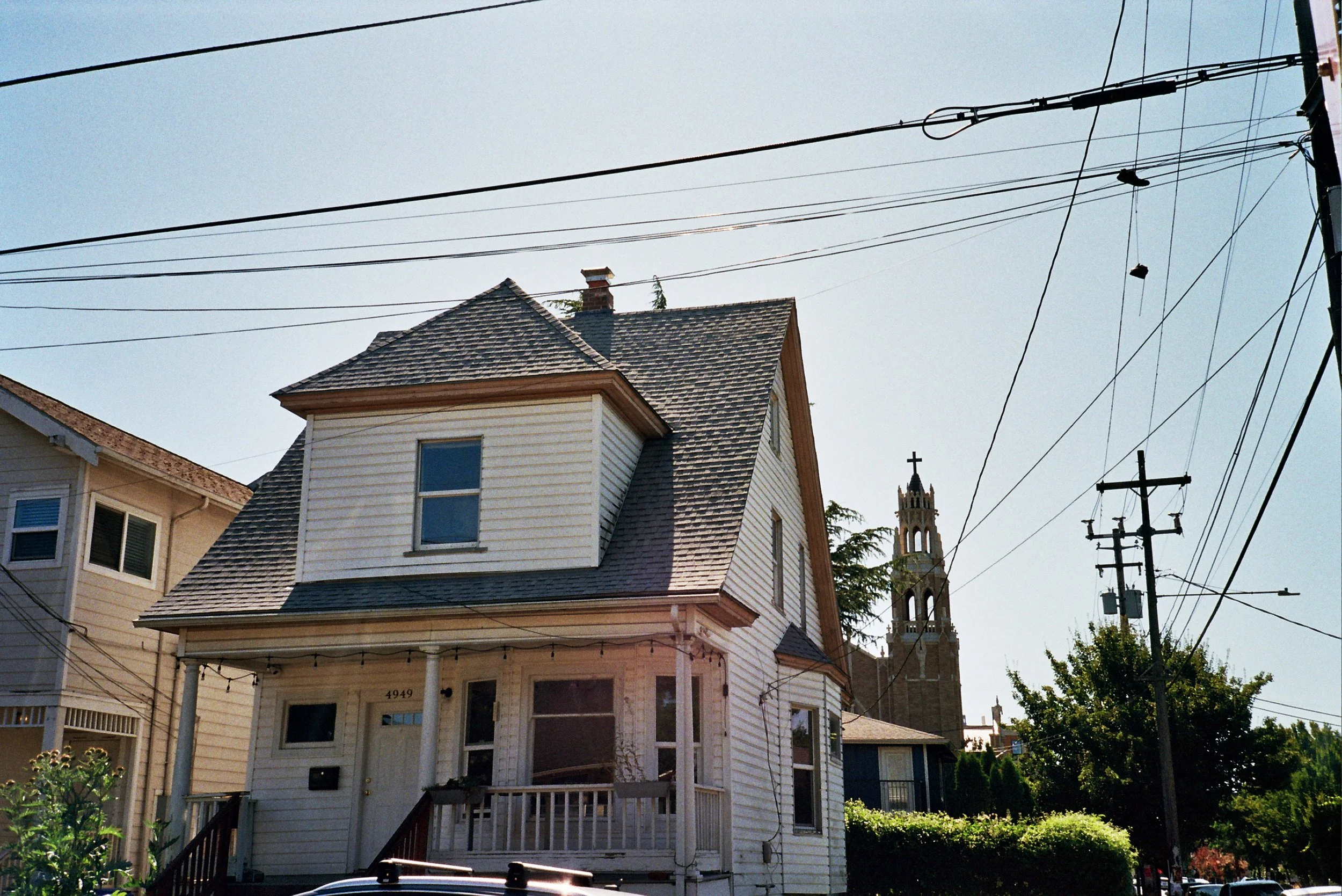 A two-story white house with a steep gabled roof and a small front porch, with multiple electrical wires crossing above and a church tower visible in the background.