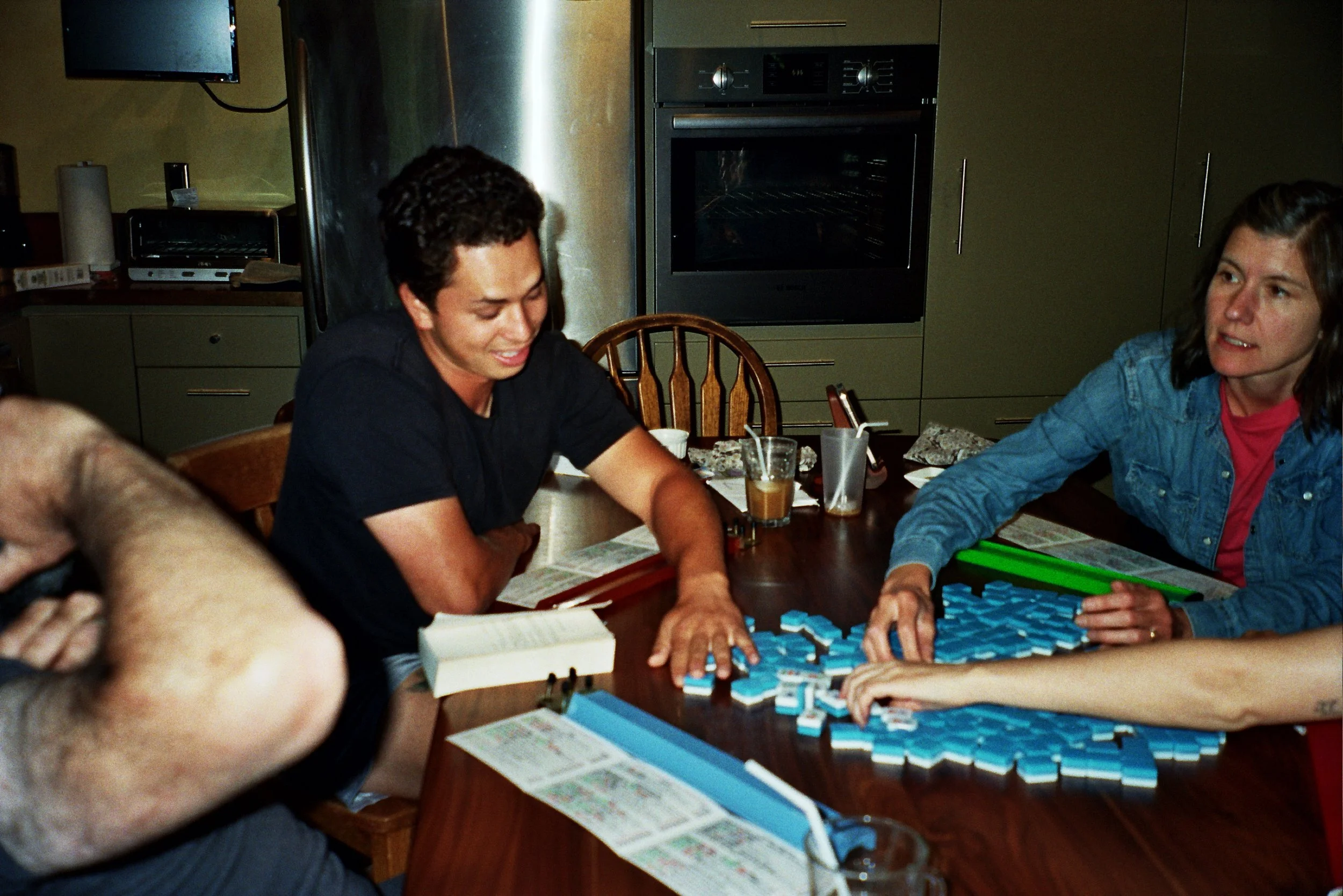 People playing a board game, possibly Scrabble, at a dining table in a kitchen. Two women are visible, one with short dark hair smiling and another with shoulder-length brown hair, wearing a denim jacket. The table has game tiles, drink glasses, and 