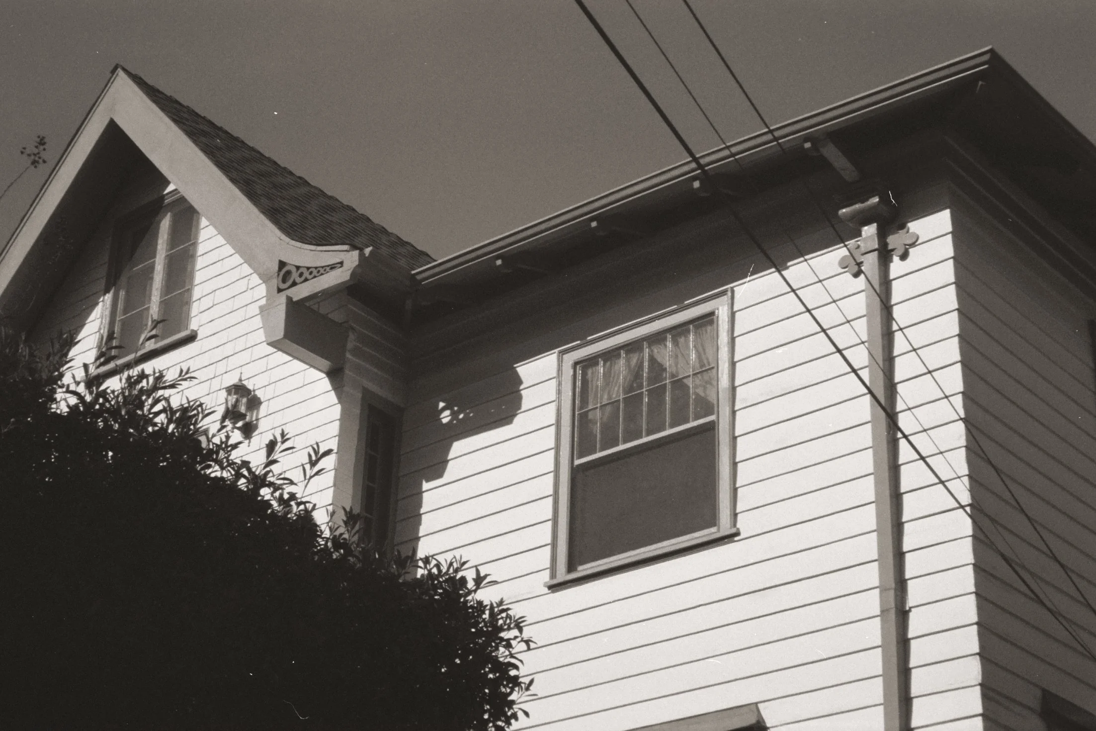 A black-and-white photo of a house with a gabled roof, windows, and an exterior light fixture, seen from below with power lines crossing in front.