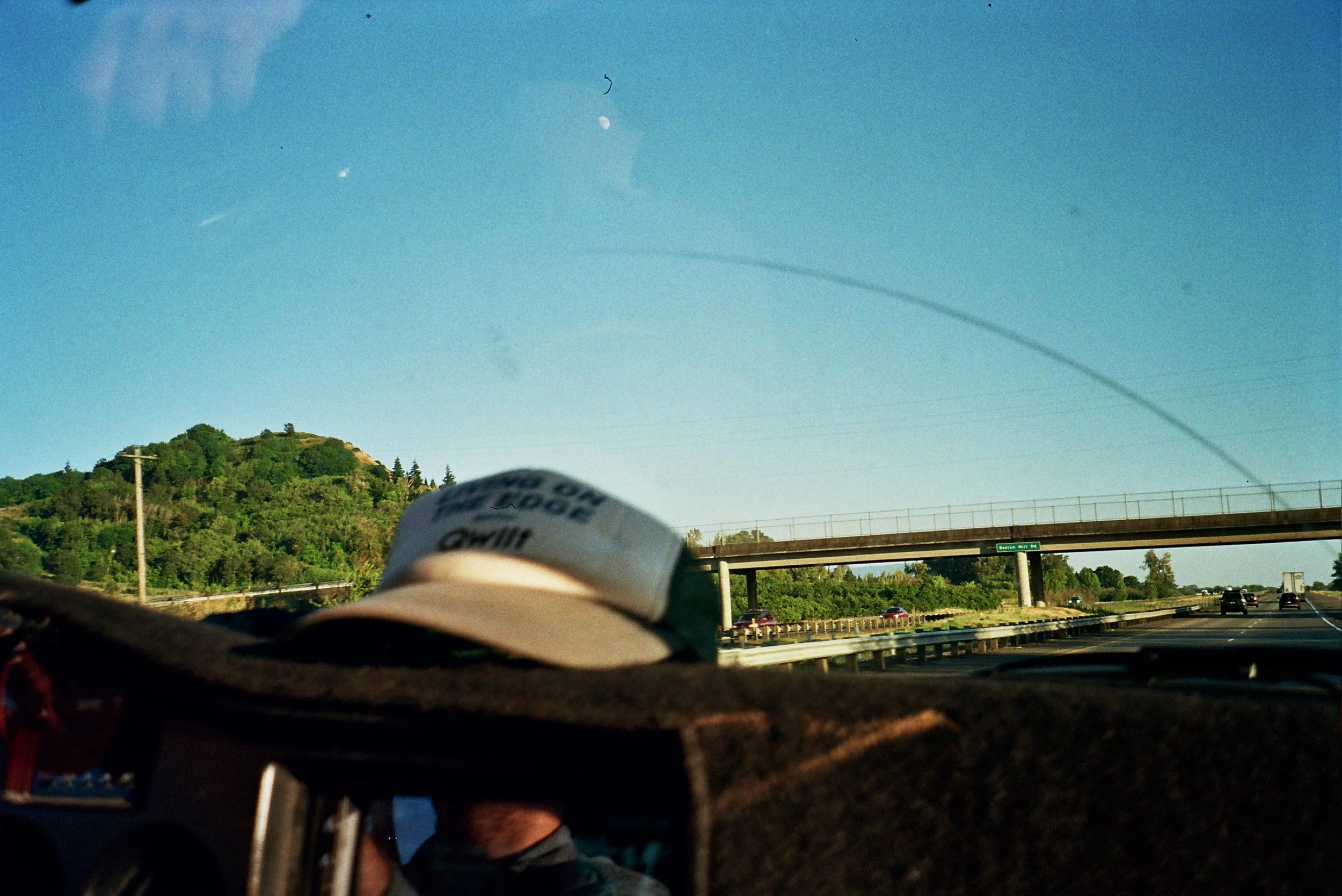 Photo taken from a moving vehicle showing the sky with the moon, a hill with trees, and a highway with cars. Coffee cup in the foreground.