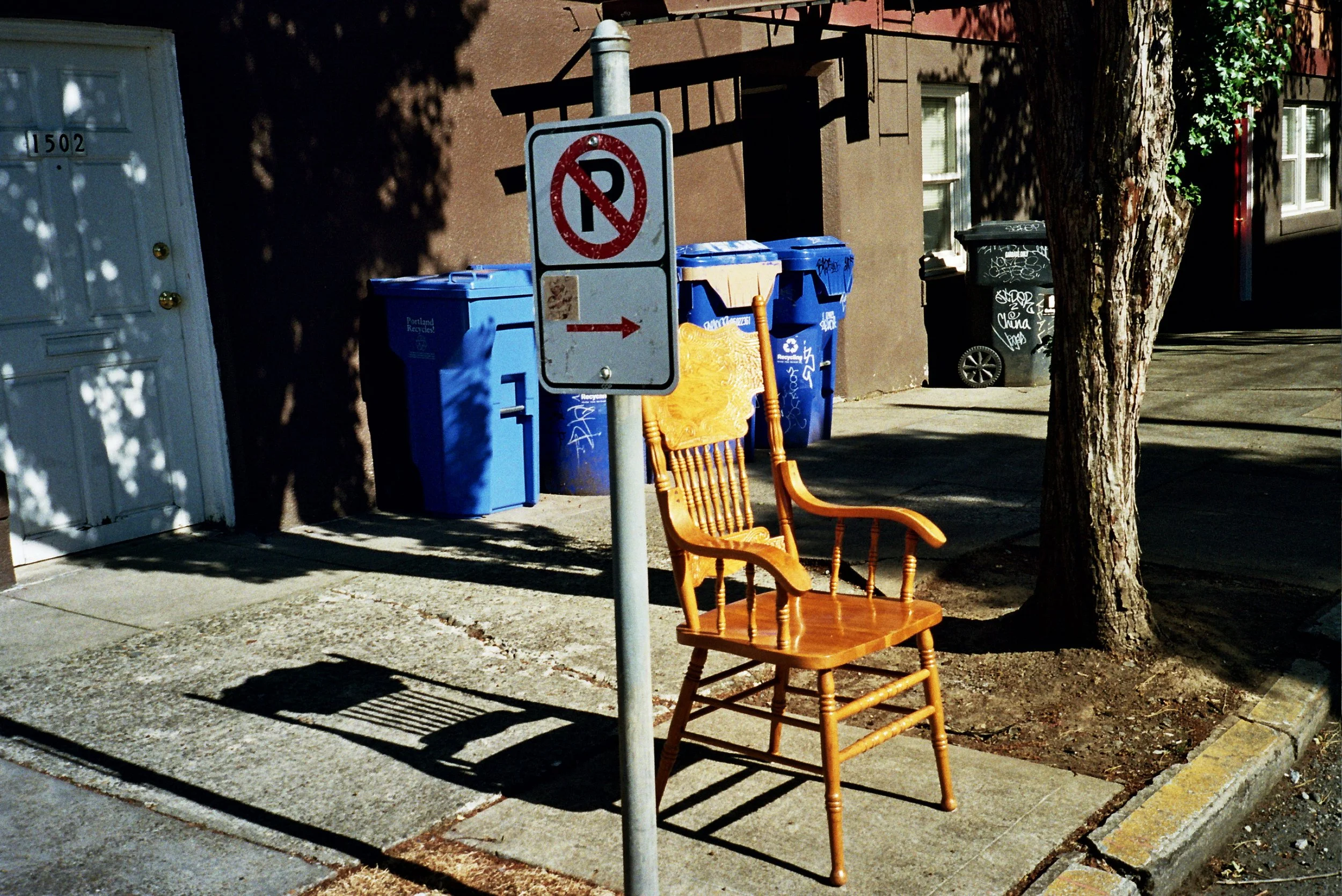 A sidewalk scene with two orange wooden chairs, a tree, and a pole with a no parking sign. Several trash bins are in the background near a brown building.