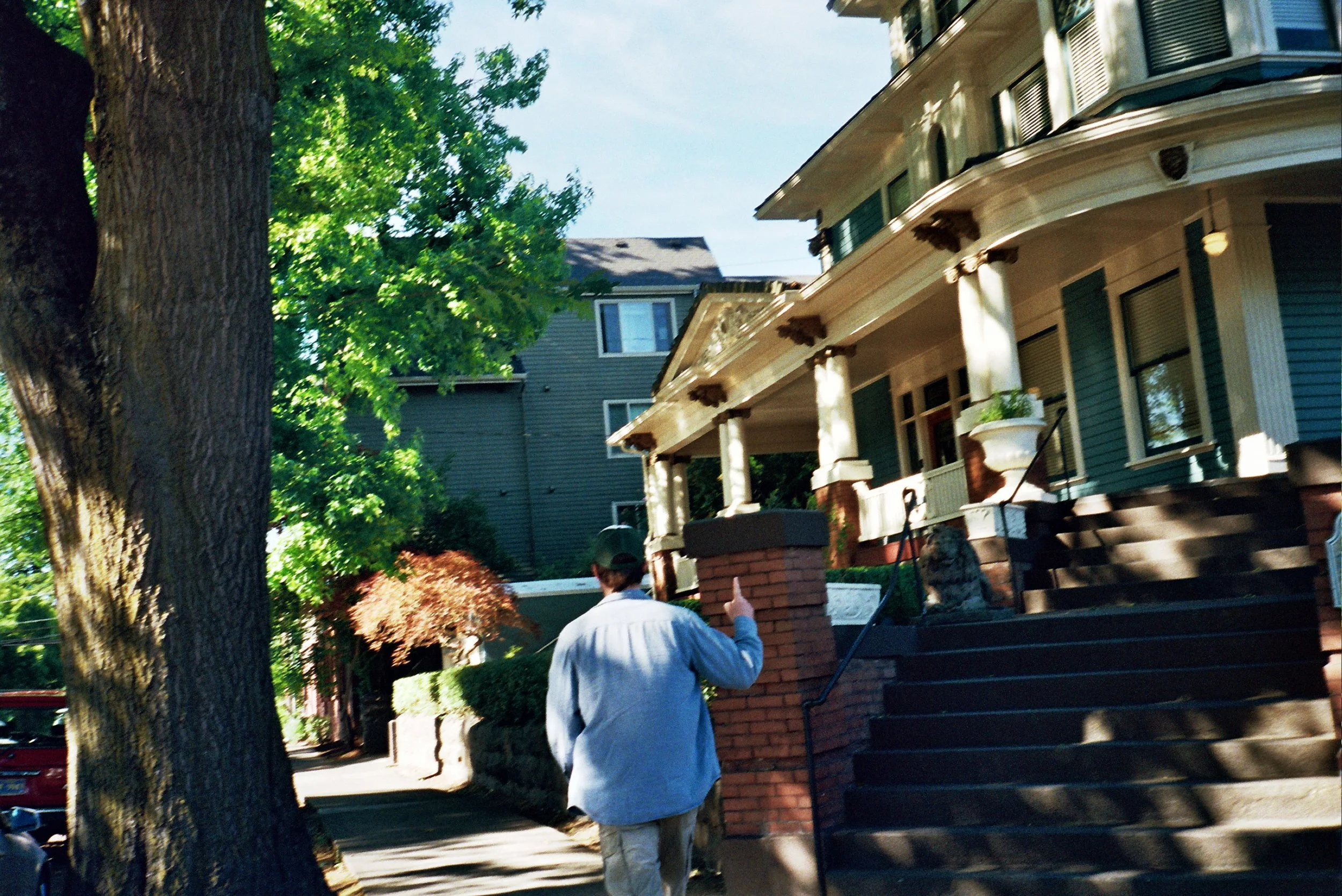Man in a white shirt walking on a sidewalk near a wooden fence and large trees in front of a blue house with a porch.