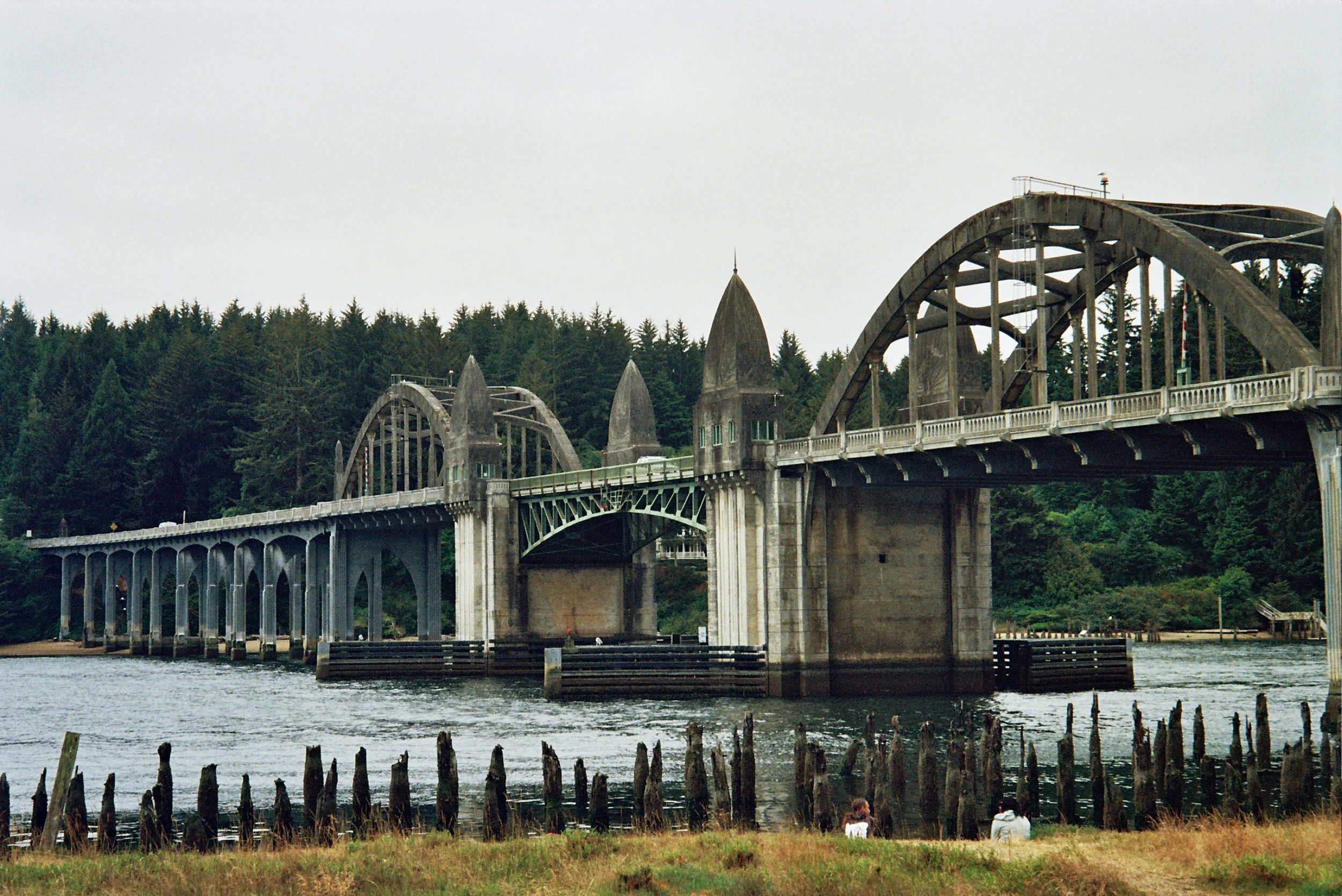 View of a large, historic bridge over a body of water, surrounded by lush green trees, with two people sitting on the grass near the water.