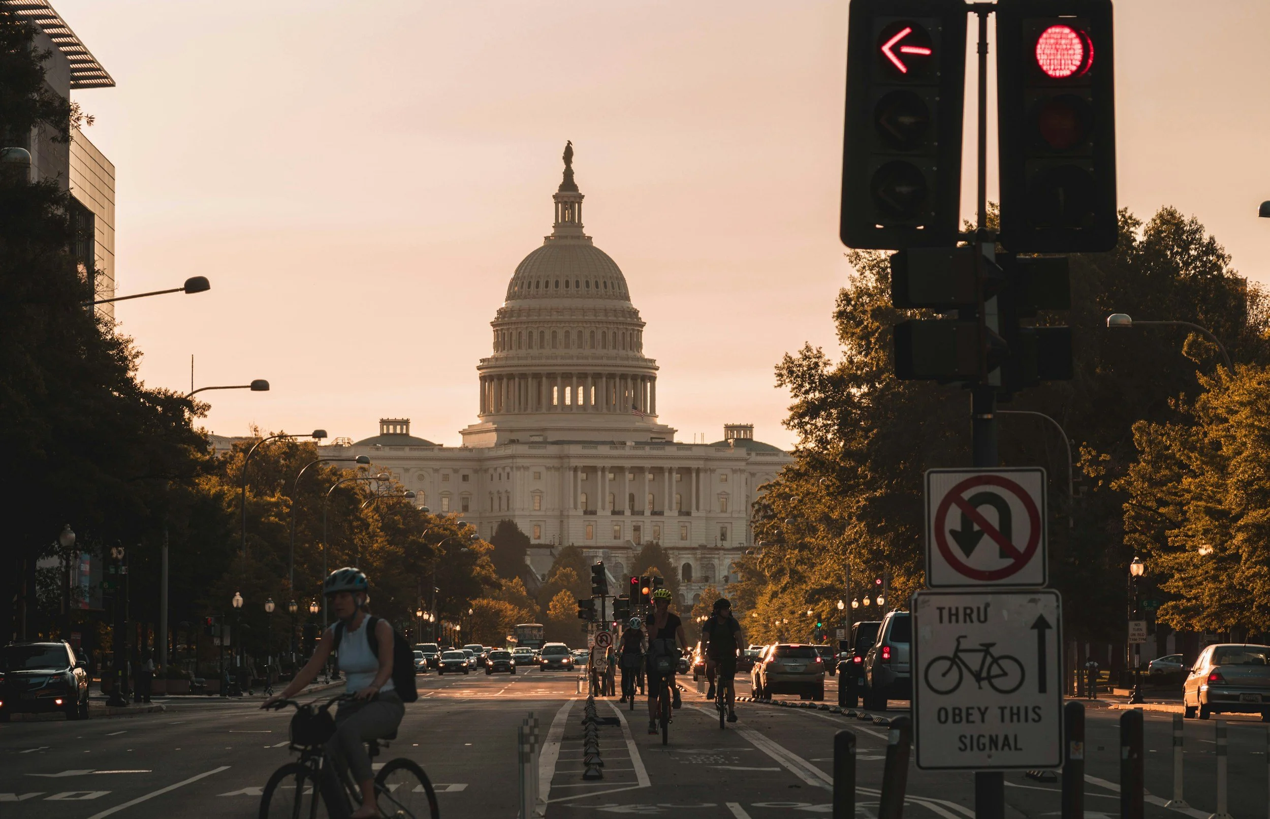 View of the United States Capitol building in Washington, D.C., during sunset with a street scene in the foreground featuring cyclists, pedestrians, and traffic signals.