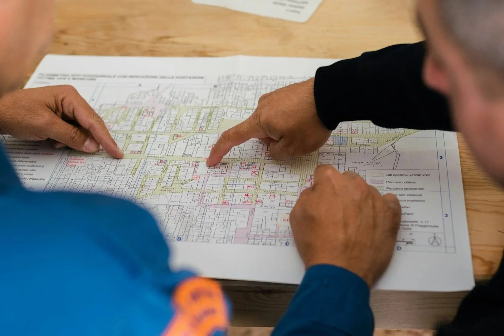 Two people are examining a detailed map or blueprint spread out on a wooden table, with one person pointing to a specific area on the map.