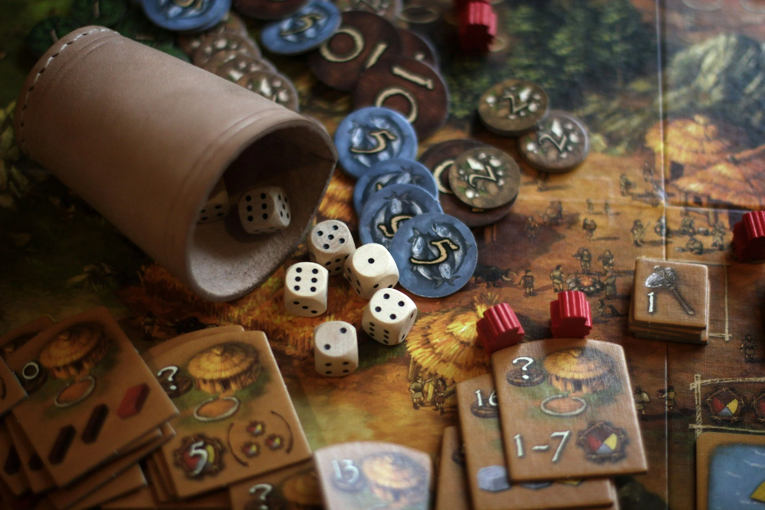 A tabletop game setup with dice spilling from a small brown cup, various colored game tokens, and cards with numbers and symbols on a game board featuring illustrations of a forest and tiny characters.