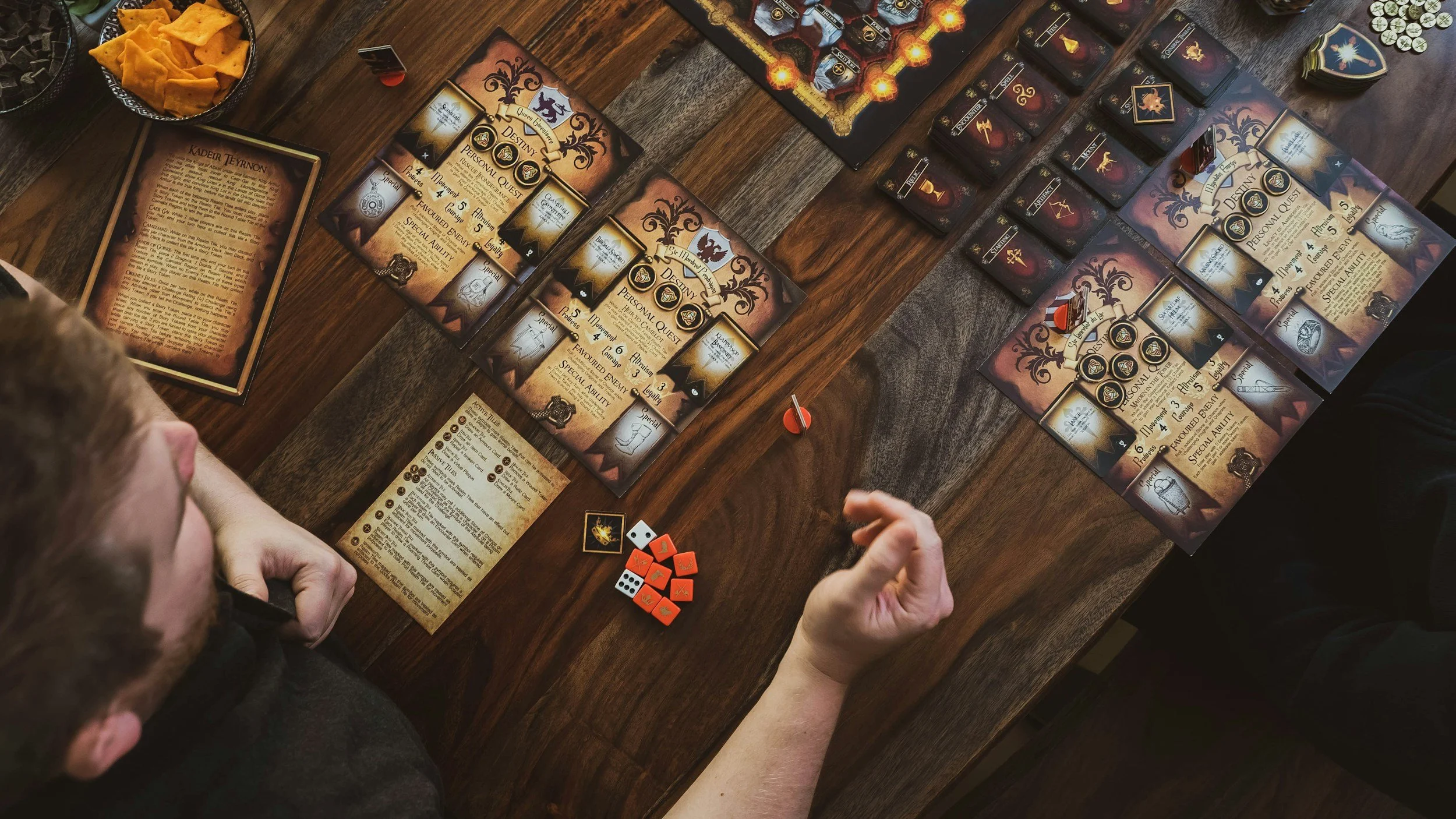 Two people playing a fantasy-themed board game at a wooden table. The game features cards, tokens, dice, and tutorial sheets, with snack bowls visible.