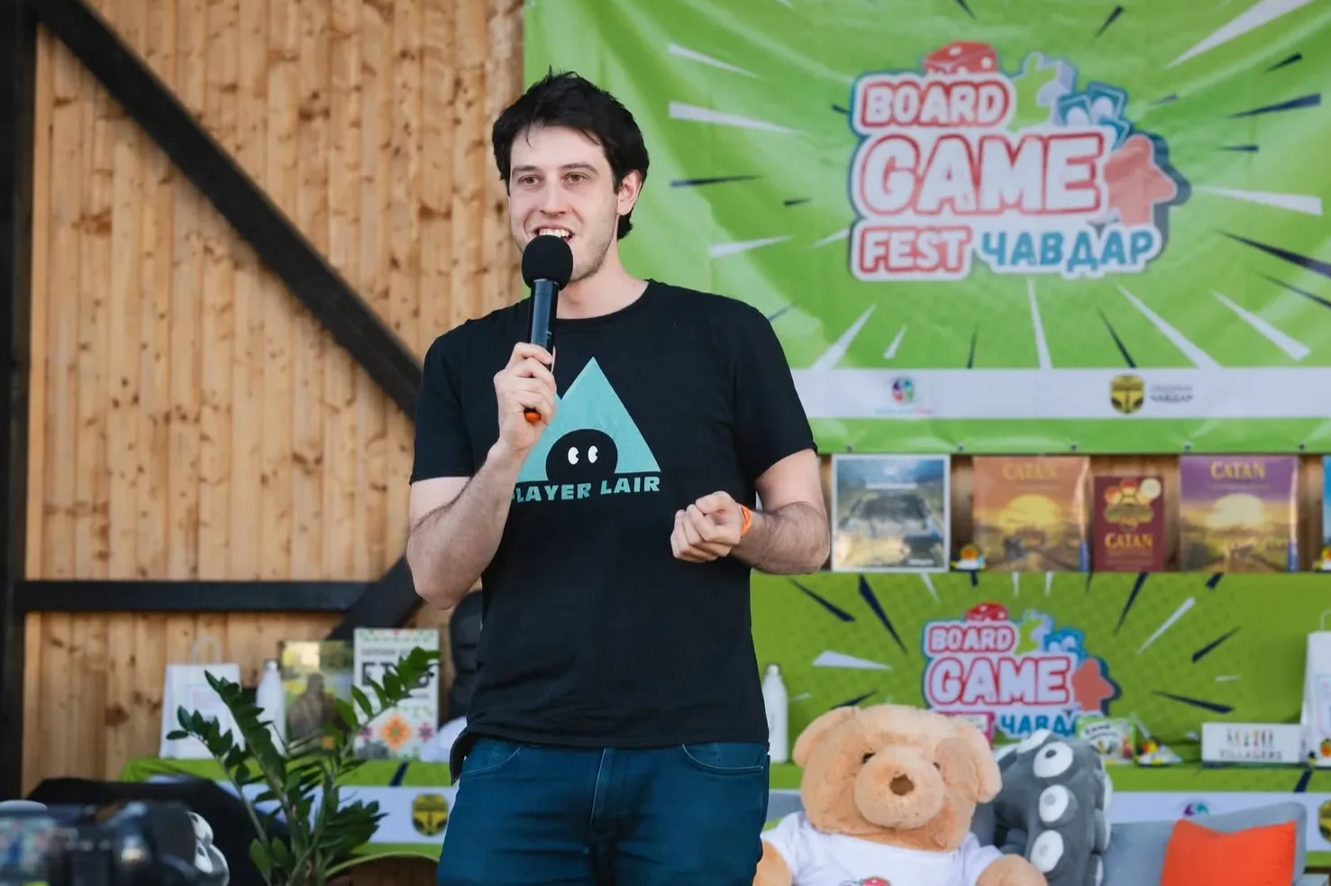 A man with dark hair and a black t-shirt holding a microphone speaks at a gaming event, with a green banner and various board games displayed in the background.