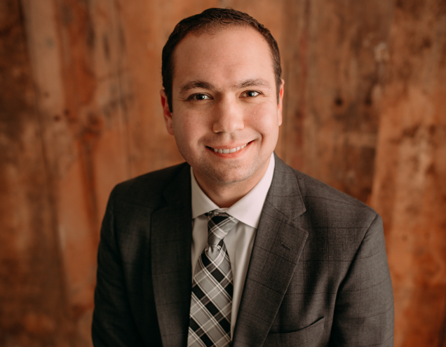 Portrait of a man in a suit and tie smiling, with a wooden background.