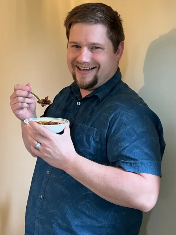 A man with short brown hair, a beard, and wearing a blue shirt, smiling, holding a bowl of food in one hand and a spoon in the other.