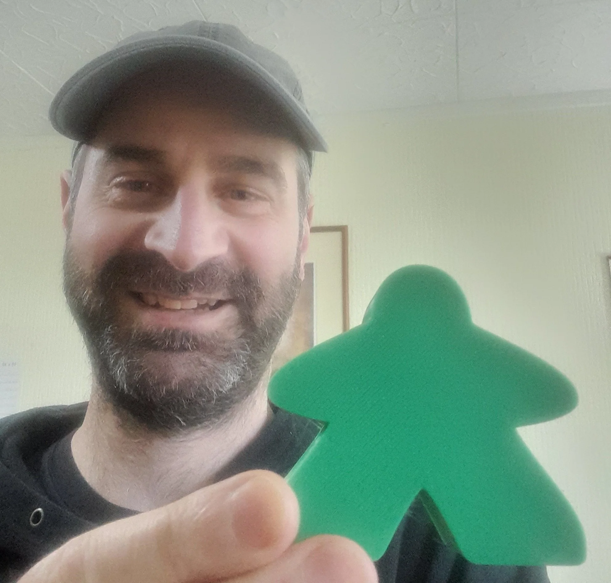 A man with a beard and mustache wearing a black cap and black hoodie, smiling and holding a green cutout of a gingerbread man, indoors with framed pictures on a light-colored wall in the background.