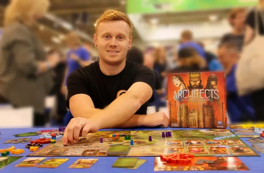 A young man with red hair and a black shirt sits at a table playing the board game 'Architects of the West Kingdom.' The game components, including cards and colorful tokens, are spread out on the table. In the background, there are other people in a busy, indoor setting.
