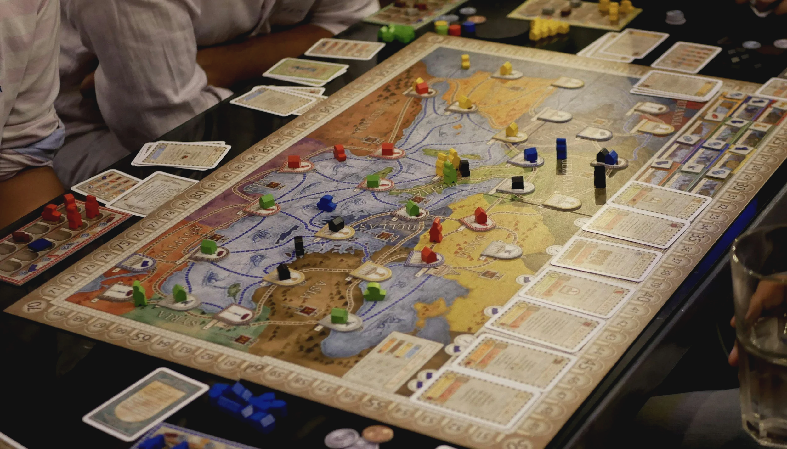 A close-up of a strategic board game in progress with colorful game pieces, cards, and a map of Europe on the board, surrounded by players' hands and drinks.