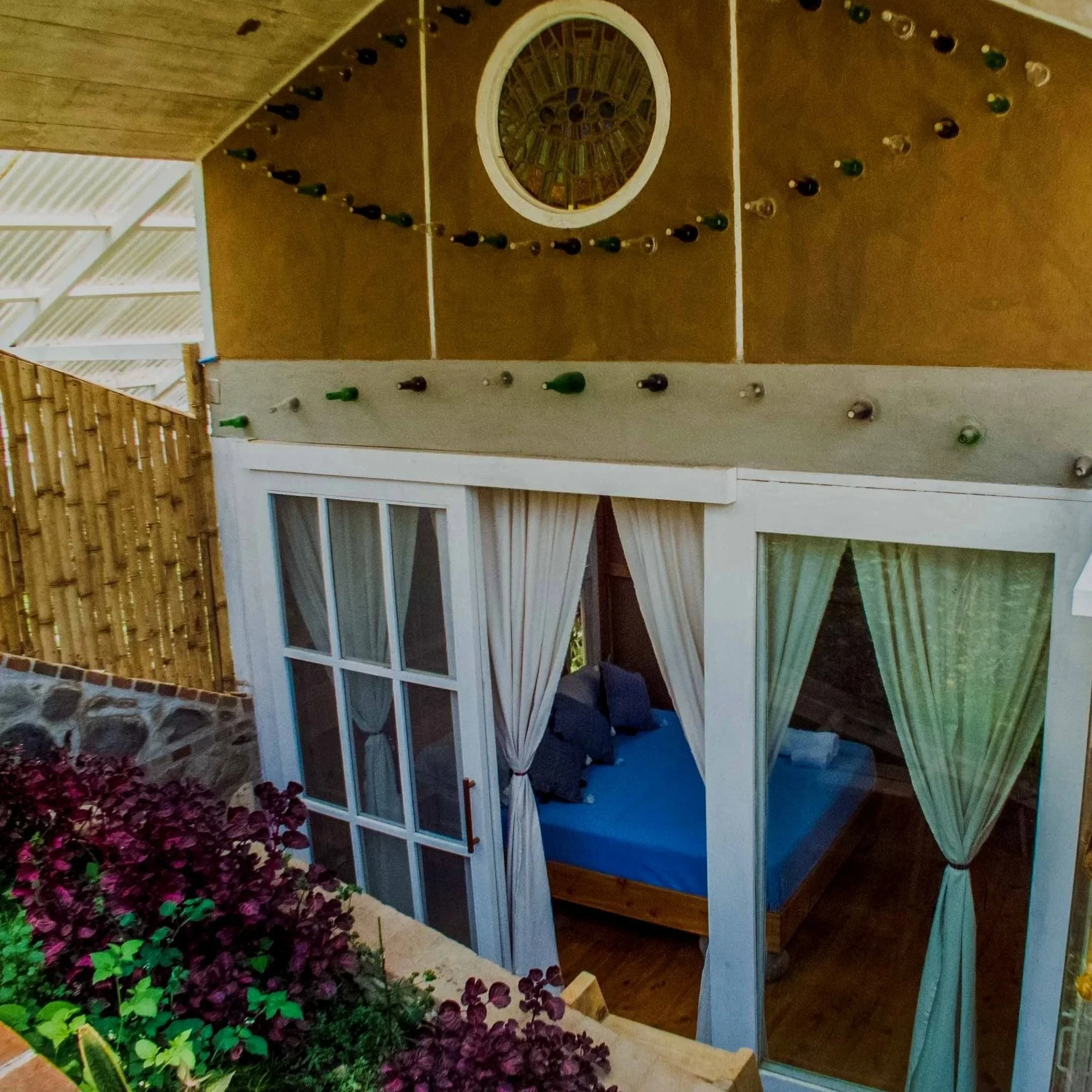 Jardín con plantas de color morado y verdes, y una estructura de madera con ventanas y una puerta de arco decorativa.