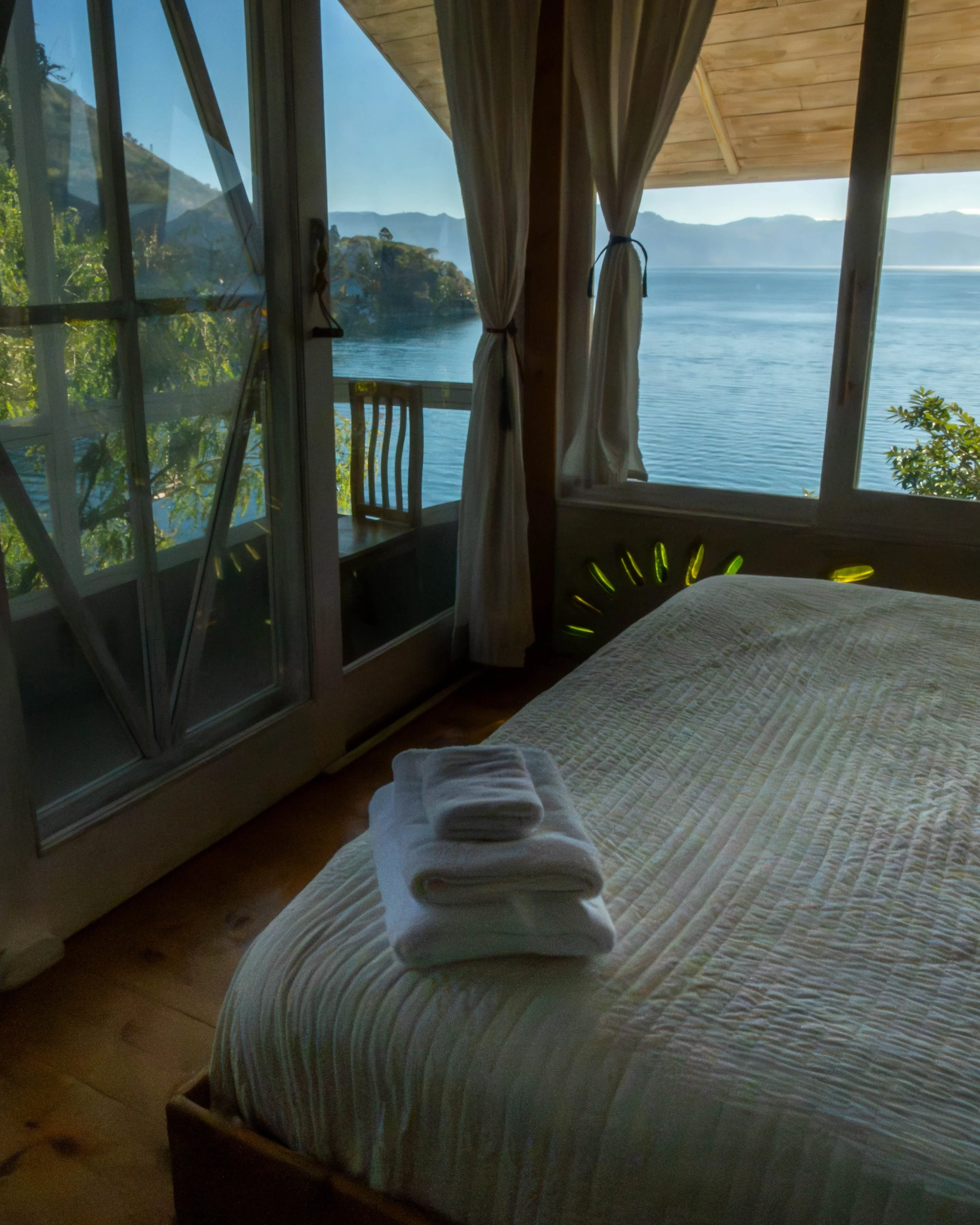 Interior de una habitación con vista a un cuerpo de agua y montañas, con una cama en primer plano y ventanas con cortinas.