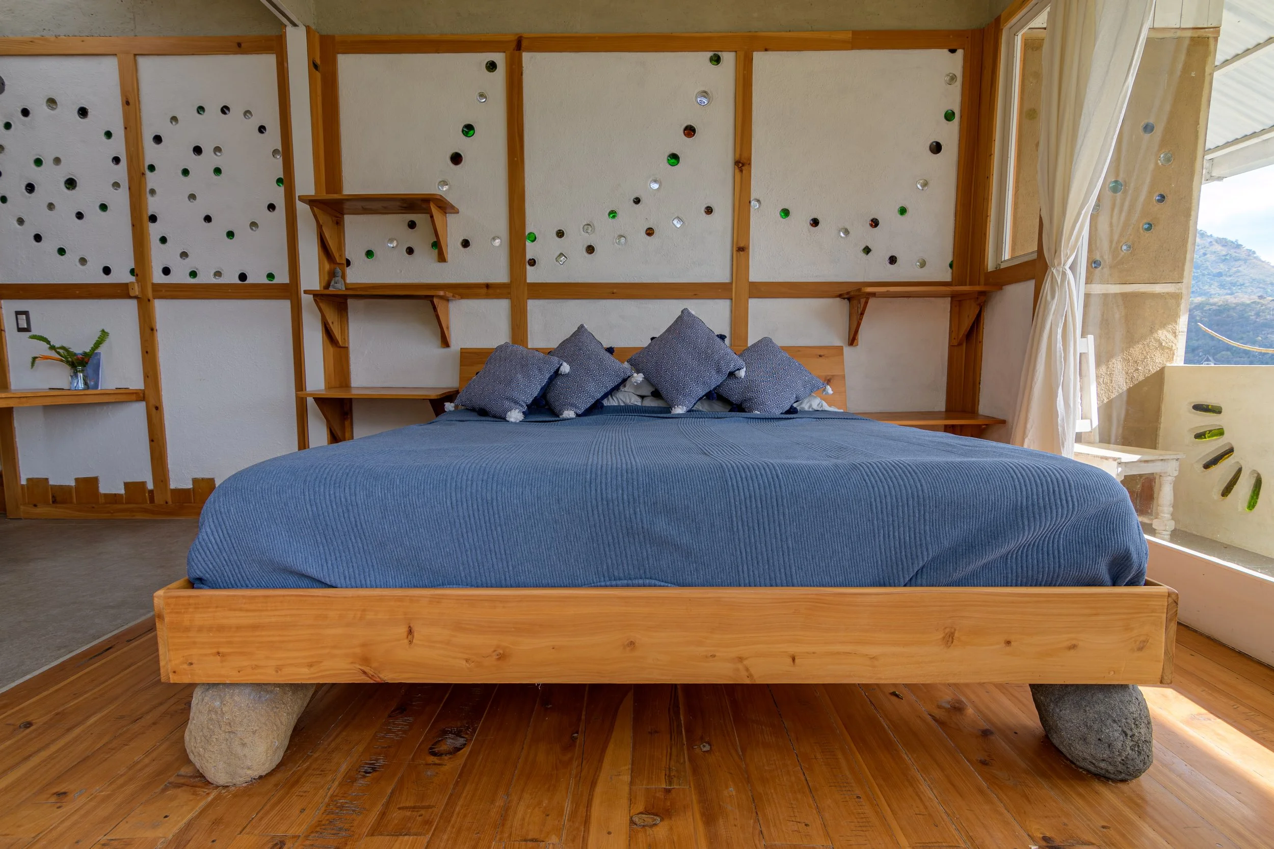 Habitación con pared tierra y madera, ventanas abiertas y cortinas blancas con vista a montañas.