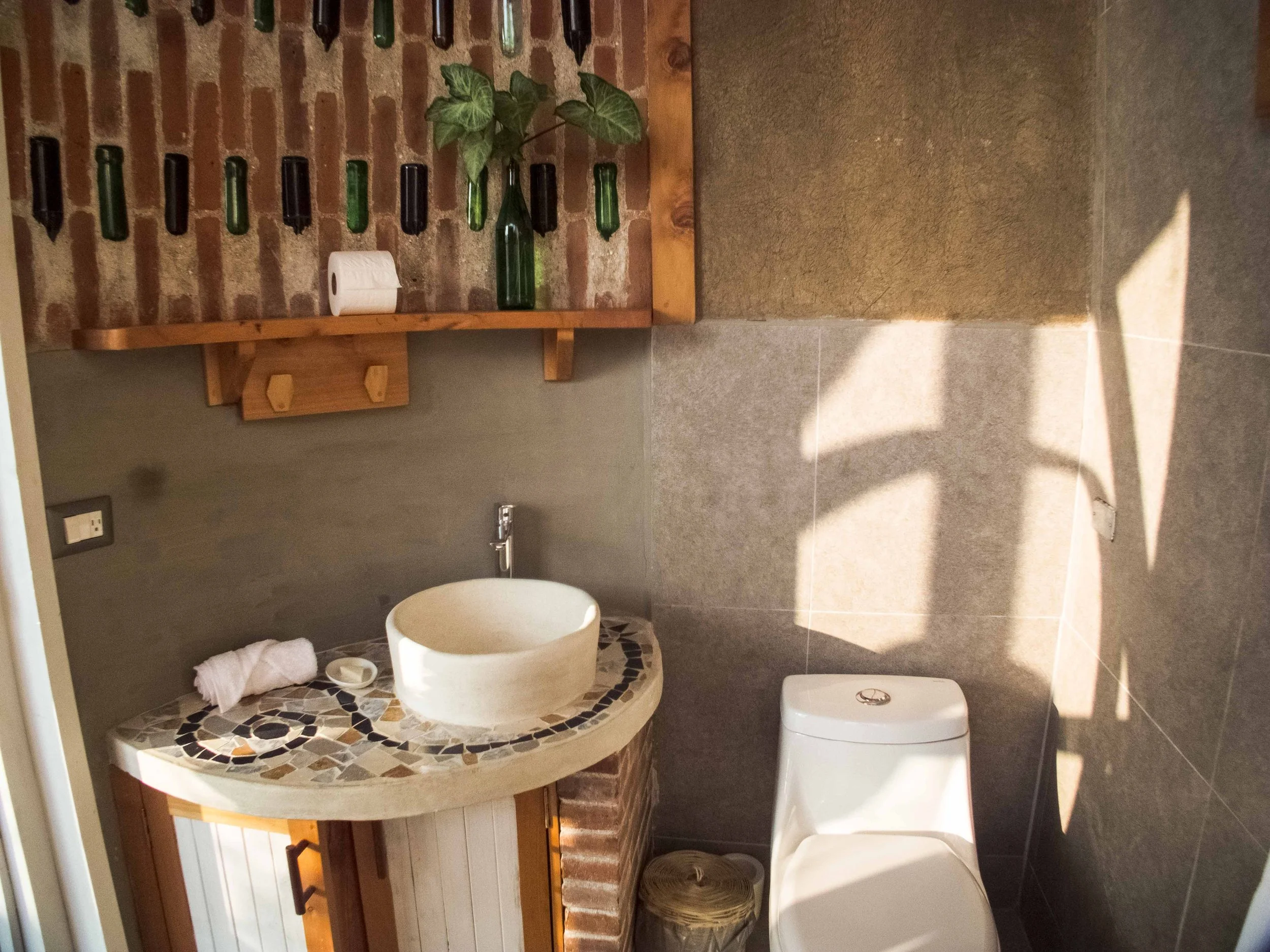 Baño con pared de ladrillo y accesorios decorativos de botellas verdes y transparentes, un lavabo de piedra con un mosaico de colores, un inodoro blanco, una papelera de mimbre, y una planta en una botella de vidrio