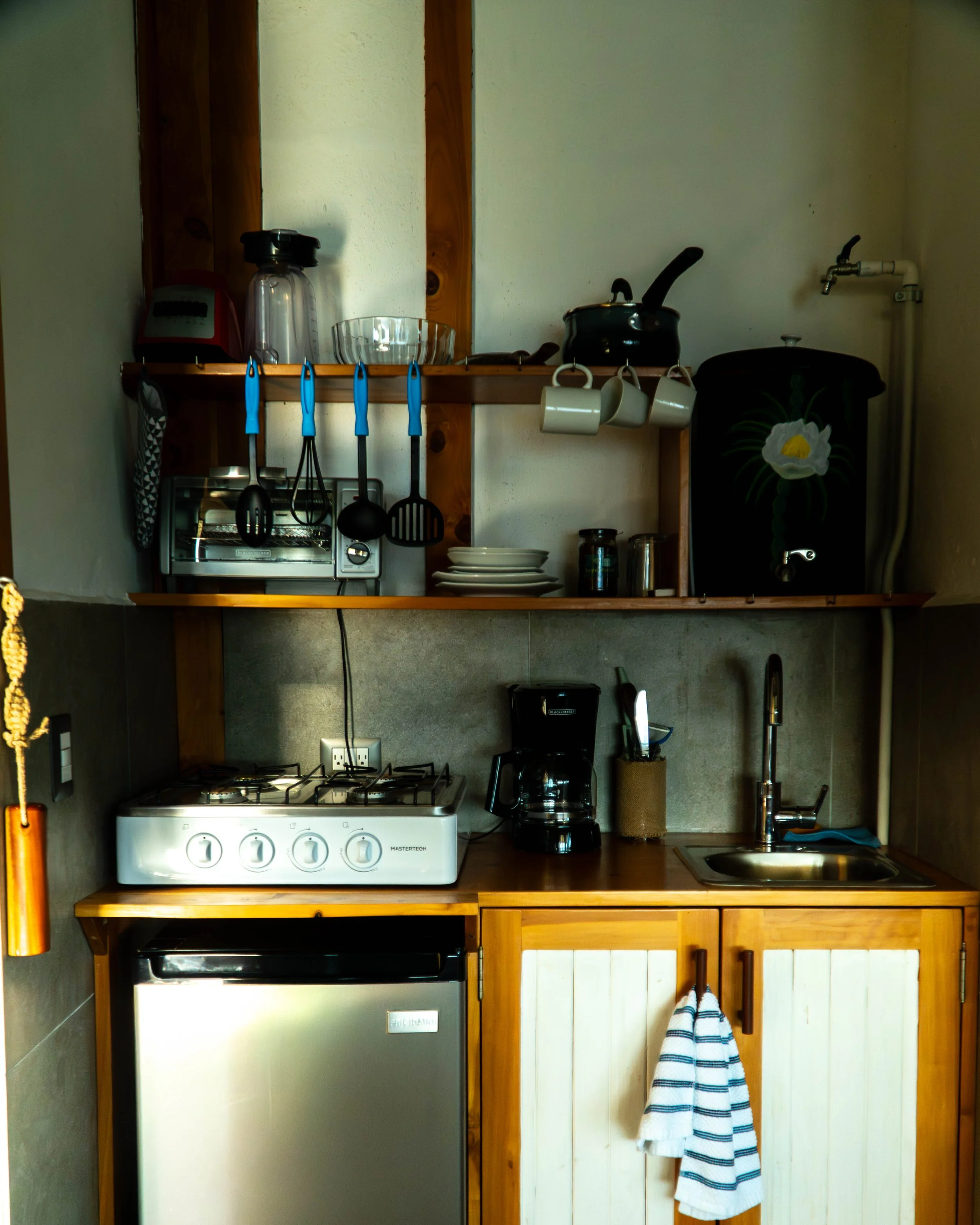 Cocina con estantes de madera, utensilios de cocina colgados, electrodomésticos como horno, cafetera, y microondas, además de tazas blancas colgadas y un toalla sobre la barra.