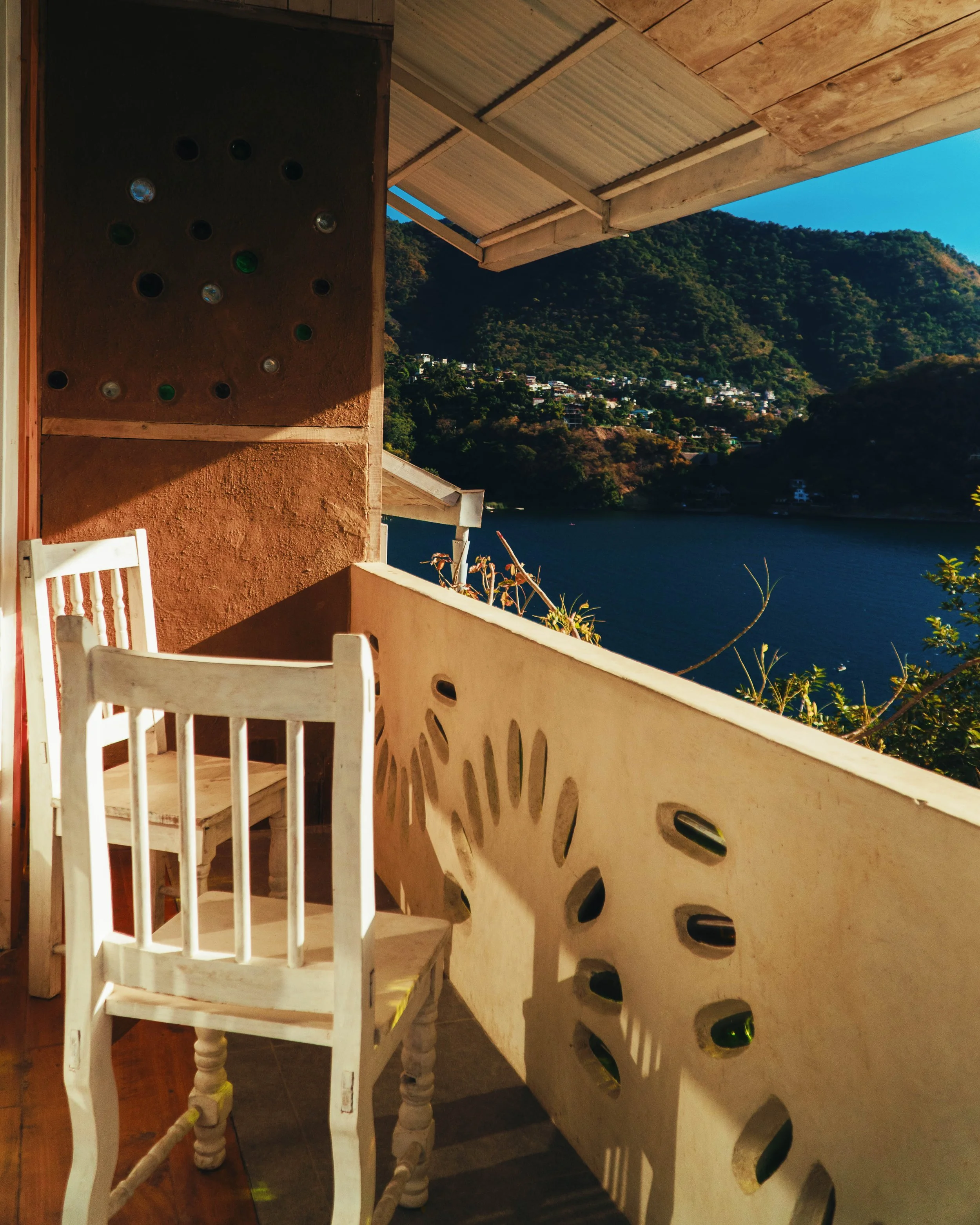 Vista desde una terraza con sillas blancas de madera, muro de color terracota con agujeros decorativos, y un paisaje montañoso con casas en la colina y un cuerpo de agua cercano, todo bajo un cielo azul claro.