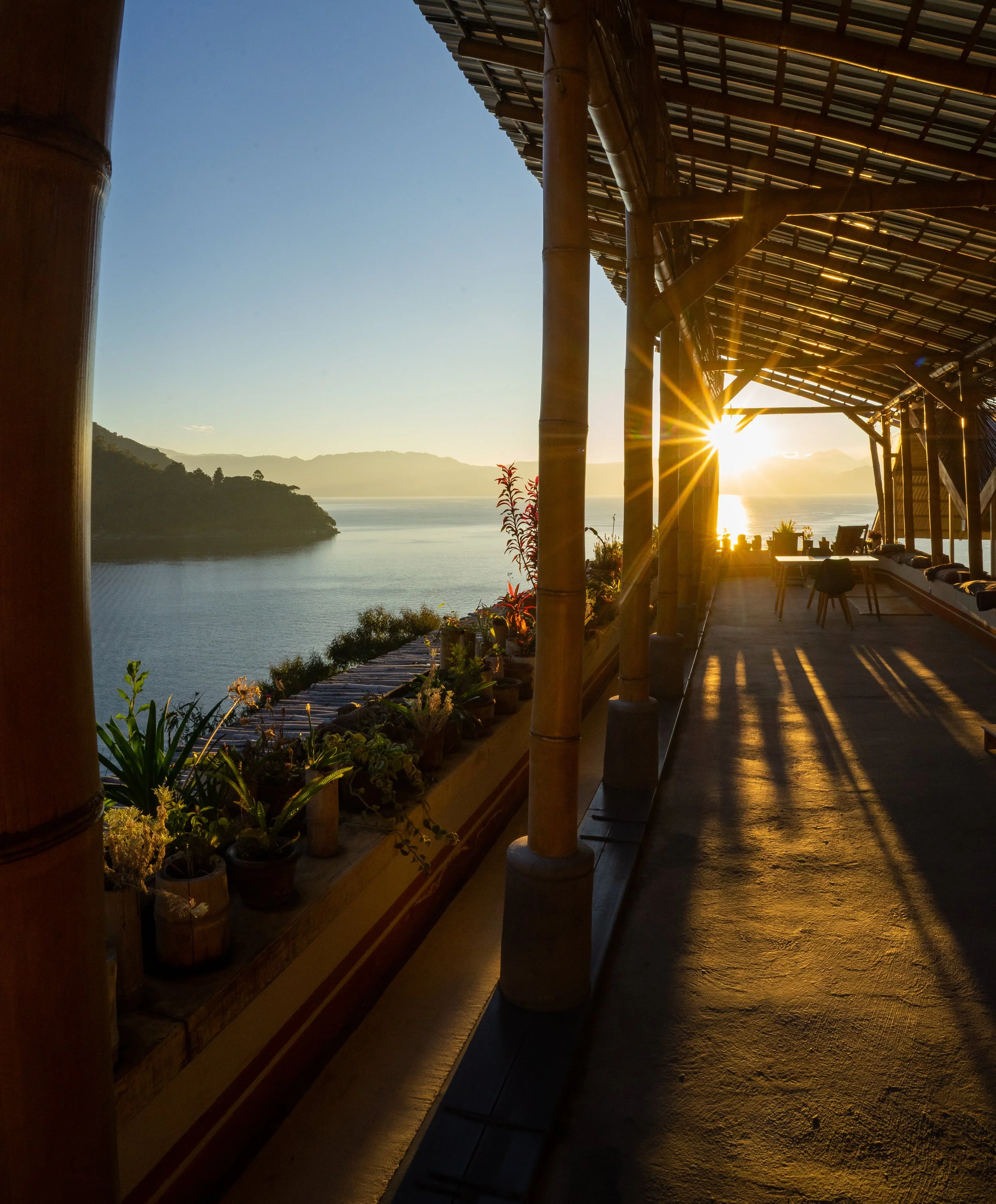 Vista del río y las montañas al atardecer desde una terraza con estructura de madera y plantas en macetas.