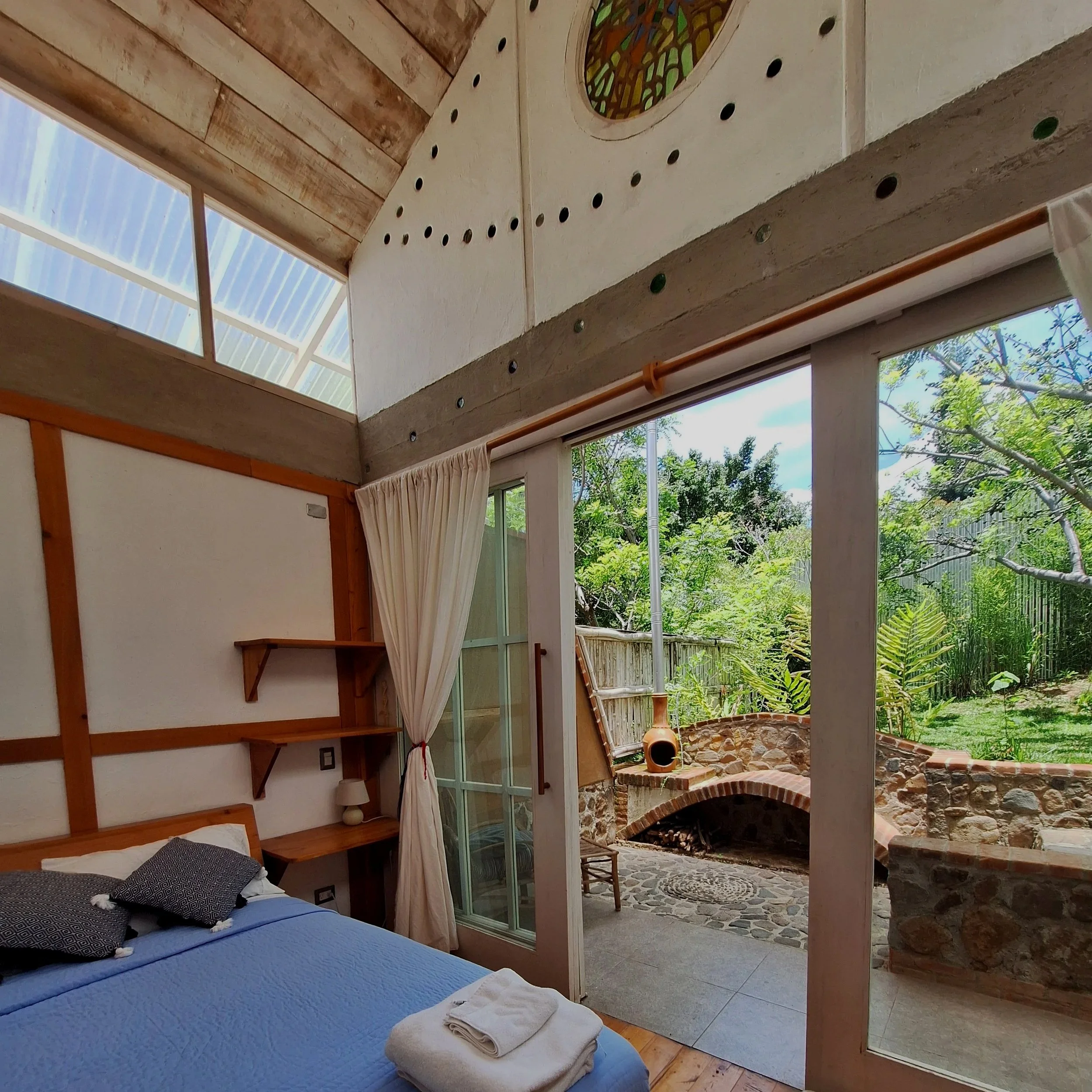 Vista desde el interior de una habitación con cama con sábanas azul claro, varias almohadas y una manta, que mira hacia una ventana grande. La habitación tiene paredes blancas, un techo de madera y un ventanal con cortinas beige. Se observa una zona exterior con césped, plantas y un horno de barro redondo en un jardín rodeado de vegetación. También hay una estructura de madera visible en el exterior.