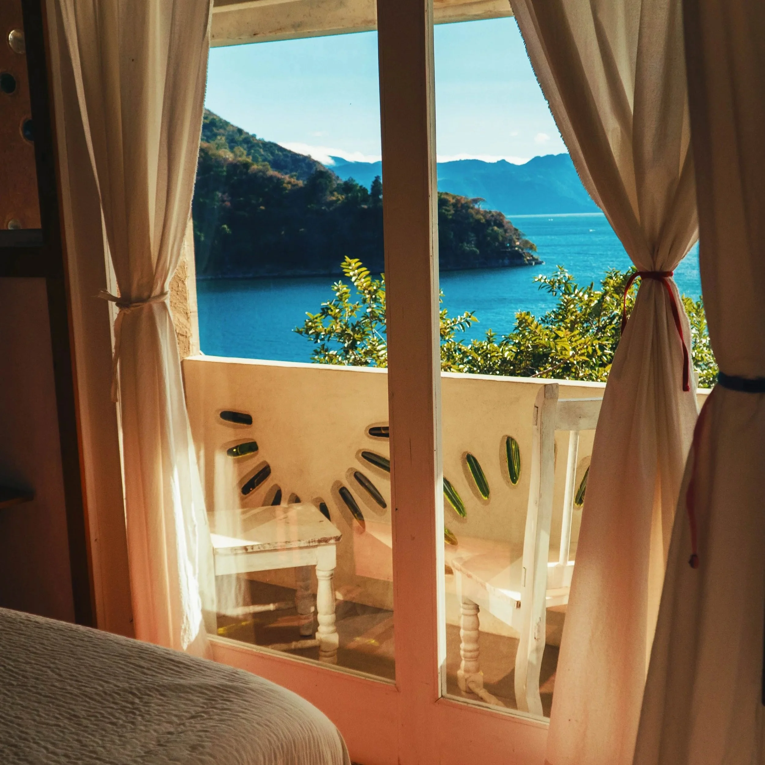 Vista de un paisaje montañoso y un lago desde una ventana con cortinas beige, en un espacio interior con paredes de tonos cálidos y muebles de madera pintados de blanco.