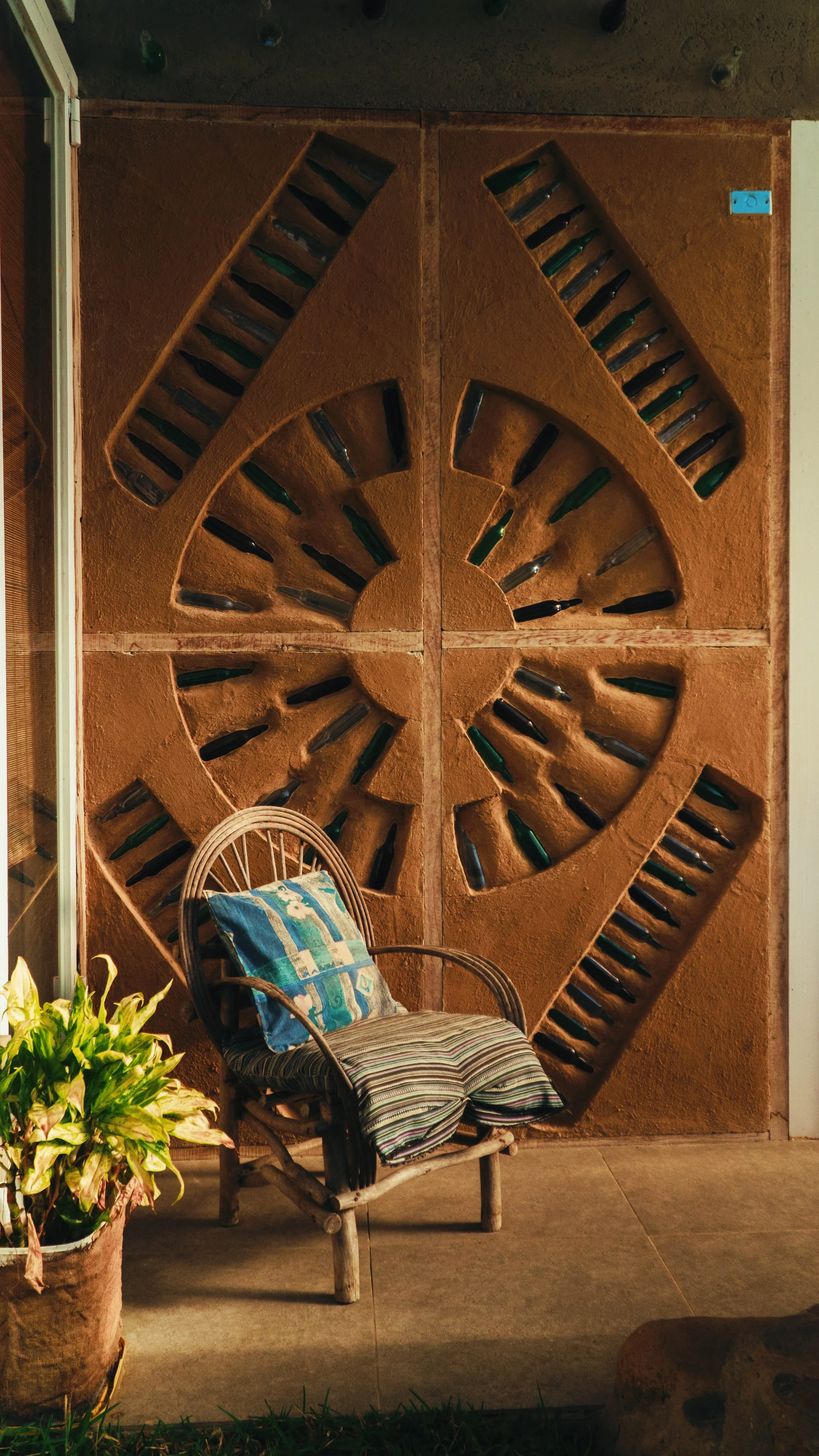 Sillón de mimbre con almohadilla de colores, plantas verdes a la izquierda y pared de ladrillos con decoraciones en forma de serigrafía de botellas de vidrio en el fondo.