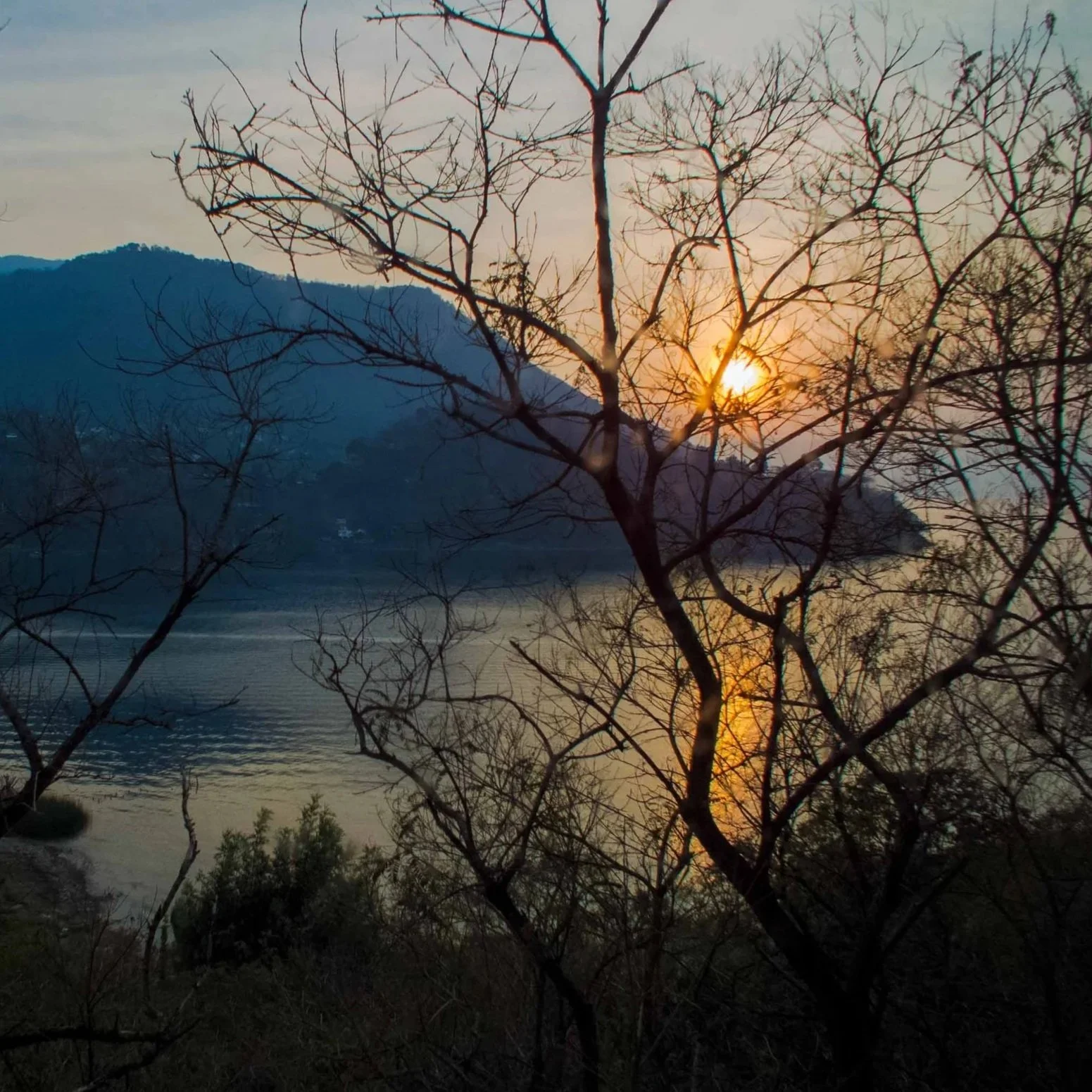 Paisaje con árboles desnudos y un río al atardecer, montañas al fondo y el sol en el horizonte.
