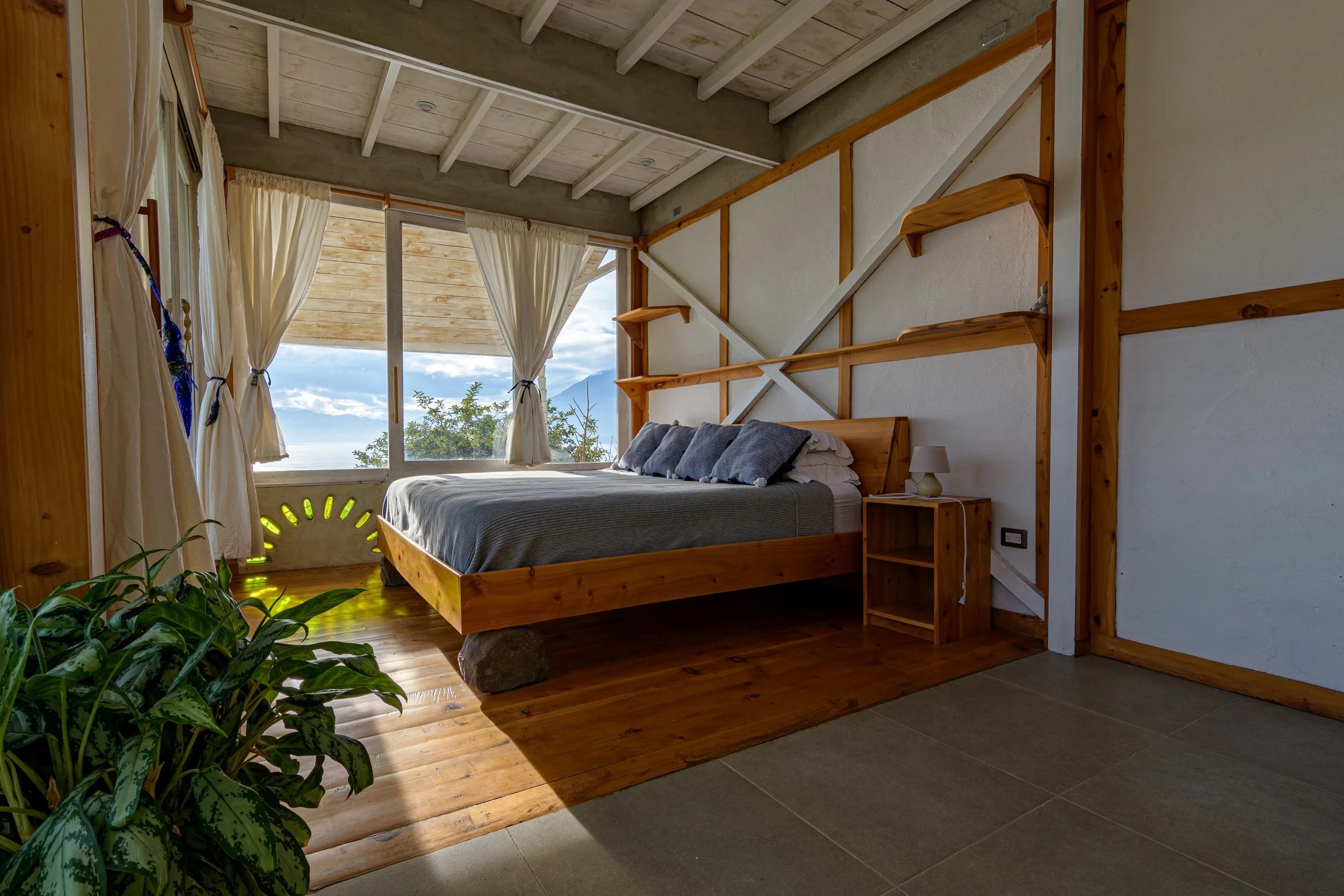 Habitación con cama de madera, varias almohadas grises, cortinas blancas, ventana grande con vista al cielo y plantas.